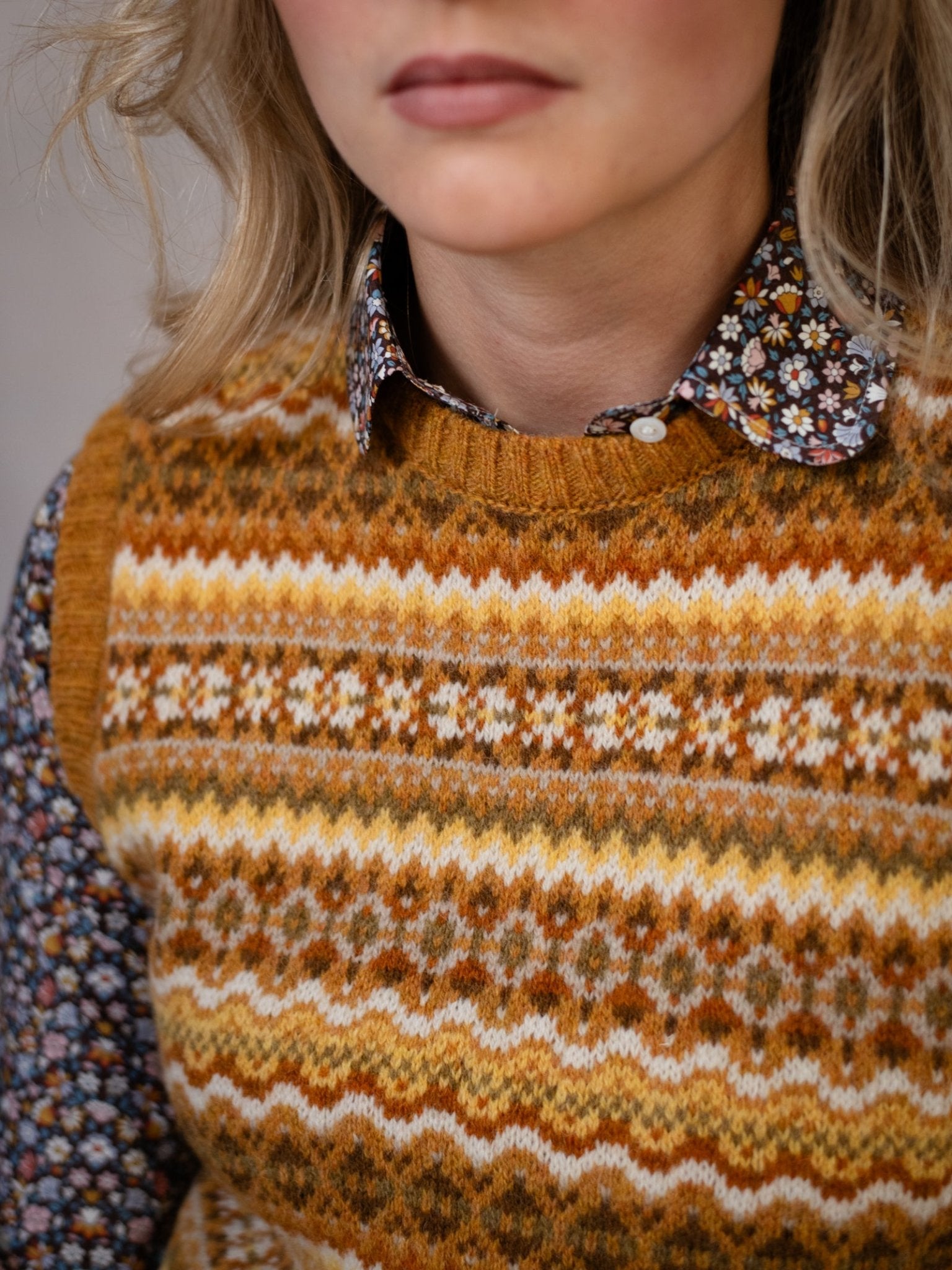 A woman wears the Campbell's of Beauly Fairisle Crew Tank, a colorful patterned knitted slipover, over a dark floral button-up shirt. The image highlights her upper torso and neck without showing her full face.