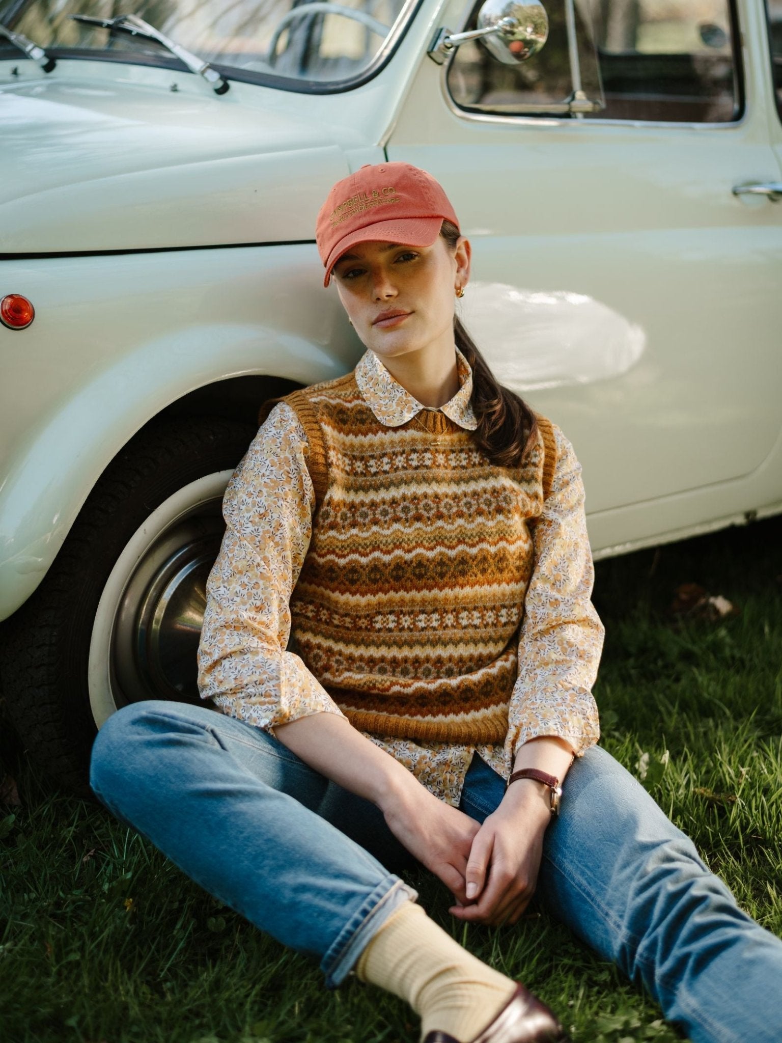 A woman wearing the Campbell's of Beauly Fairisle Crew Tank sits on grass by a white vintage car, pairing it with a floral shirt, jeans, and a pink cap, one knee up and looking relaxed.