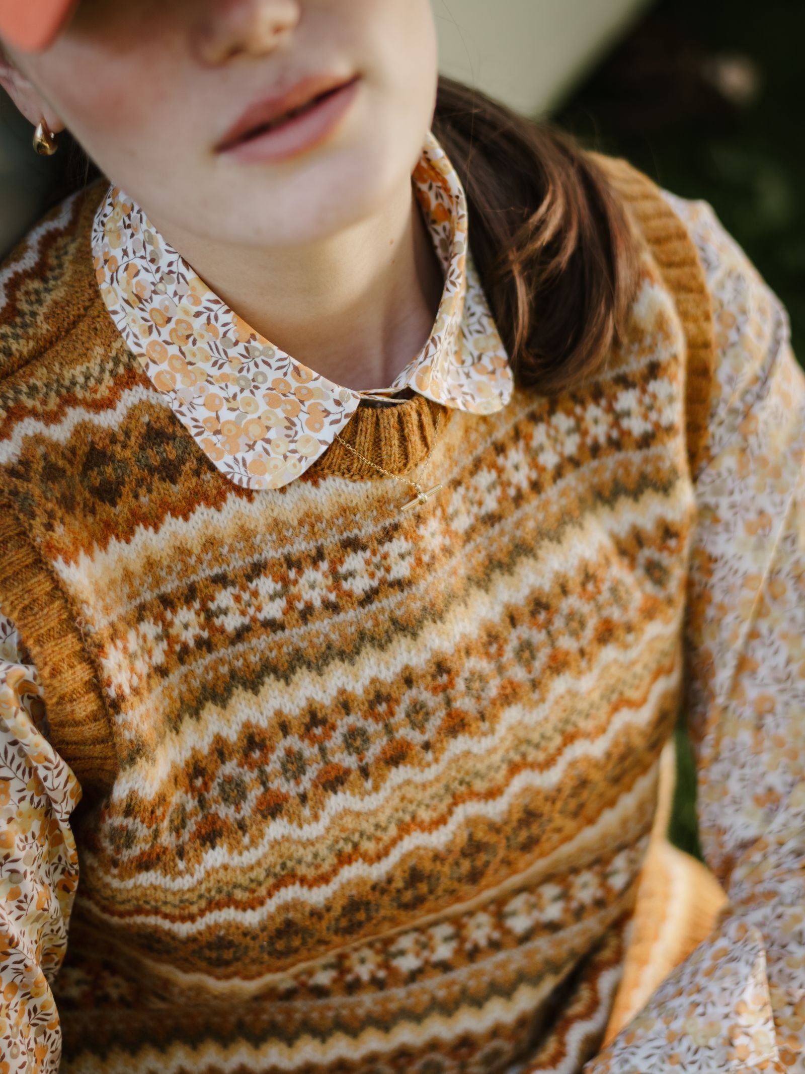A person wearing a brown patterned Campbell's of Beauly Fairisle Crew Tank, made in Scotland from Shetland wool, over a floral collared shirt and reddish cap sits outdoors. Autumnal tones and textures are highlighted; their face is partly visible.