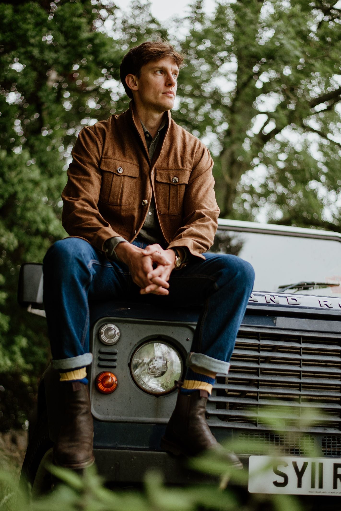A man in a Campbell's of Beauly Field Teba Jacket, blue jeans, and boots sits on the hood of a classic Land Rover parked outdoors, hands clasped as he looks to the side with trees in the background.