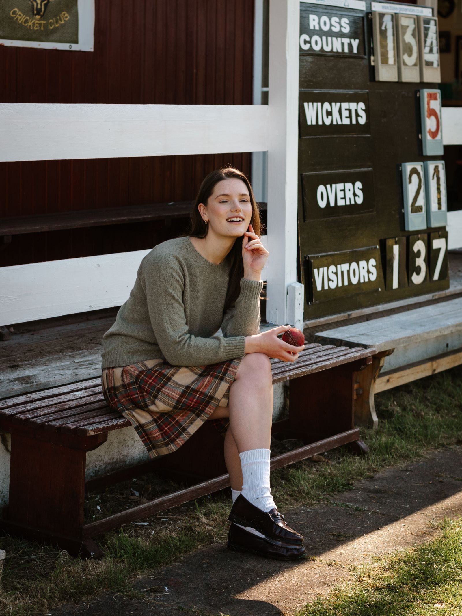 A young woman in a gray Campbell's of Beauly Geelong Lambswool Crew Neck Jumper, plaid skirt, and black shoes sits on a wooden bench holding a red cricket ball. Behind her is a scoreboard: Ross County 134/5, Visitors 137. Made in Scotland.