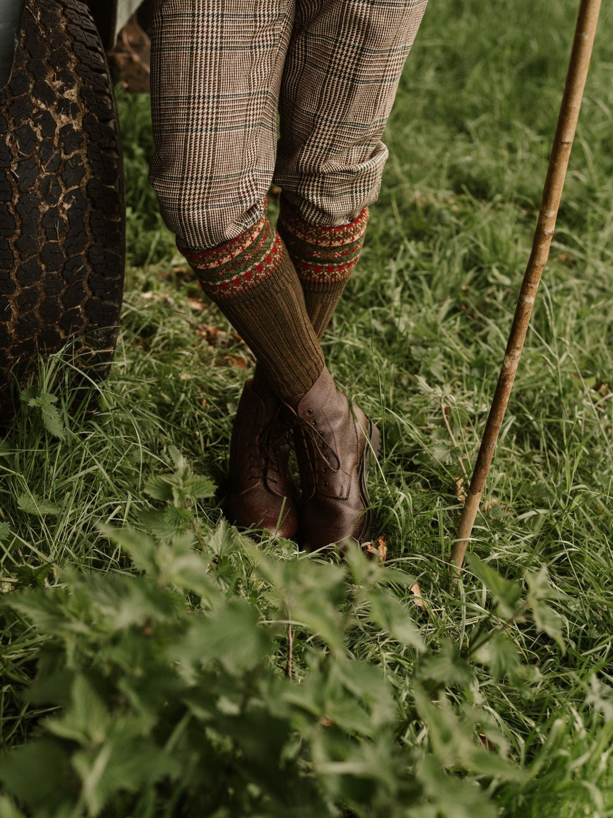 Wearing Campbell's of Beauly Tweed Breeks, classic country attire, brown boots, and patterned socks, a person stands on grass by a vehicle tire, holding a wooden walking stick.