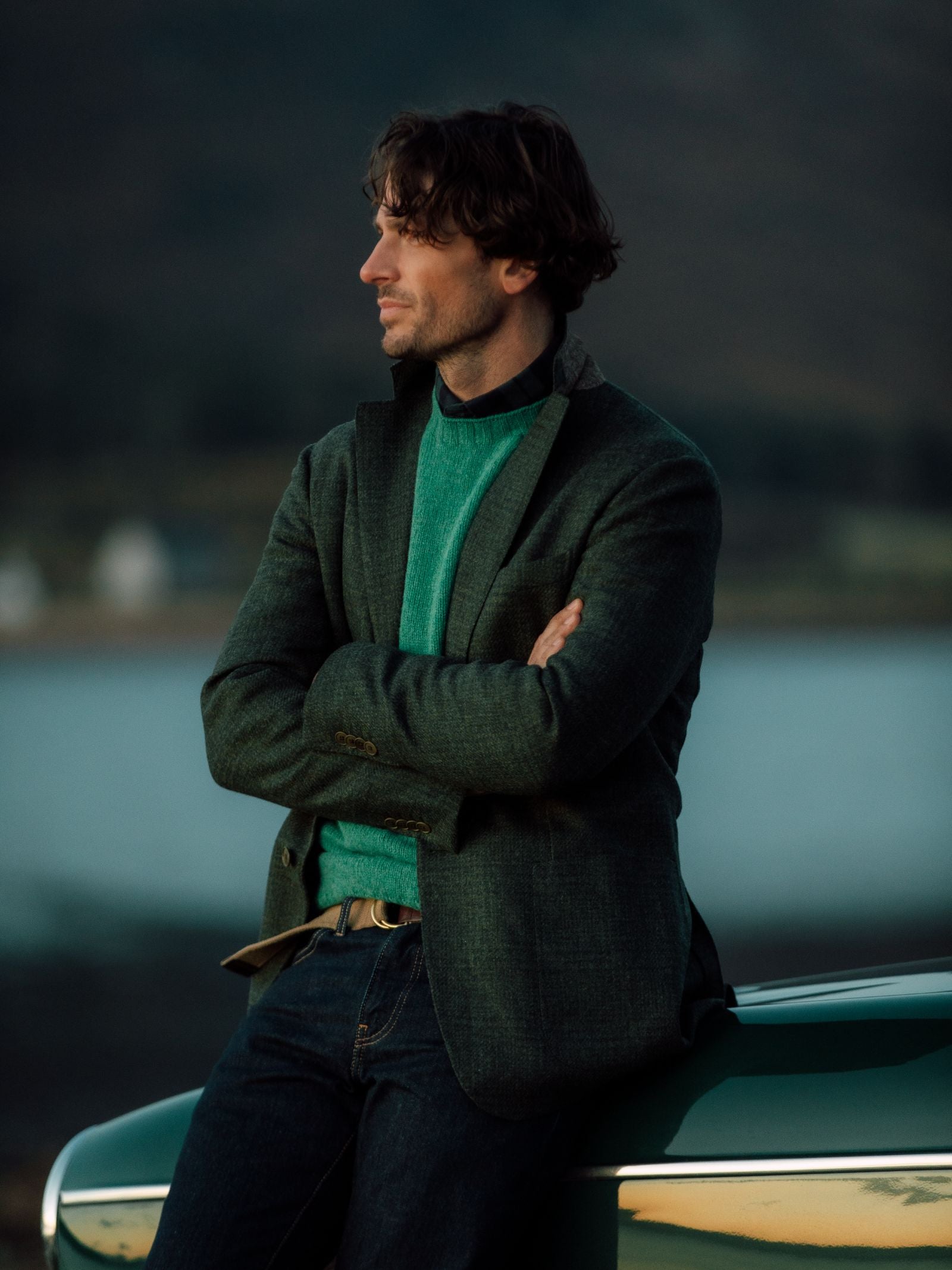A man with wavy brown hair in jeans and a Campbells of Beauly Glen Check Jacket stands by a green car near the water, arms crossed, with a blurred landscape behind him, evoking classic tailoring.
