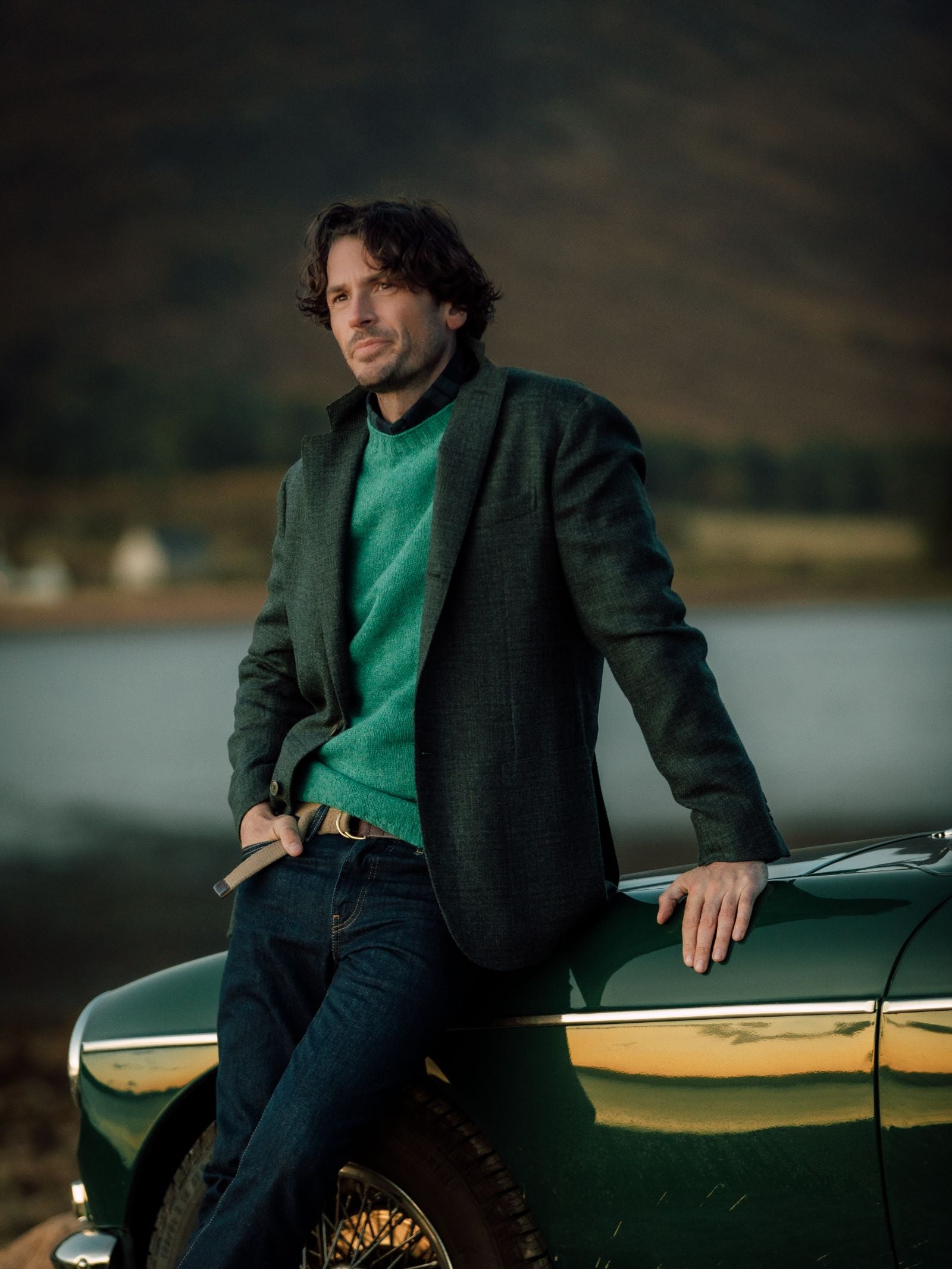 A man with wavy dark hair in a Campbells of Beauly Glen Check Jacket leans against a classic green car by a lake, hills in the background. He gazes thoughtfully into the distance, holding a cigarette.