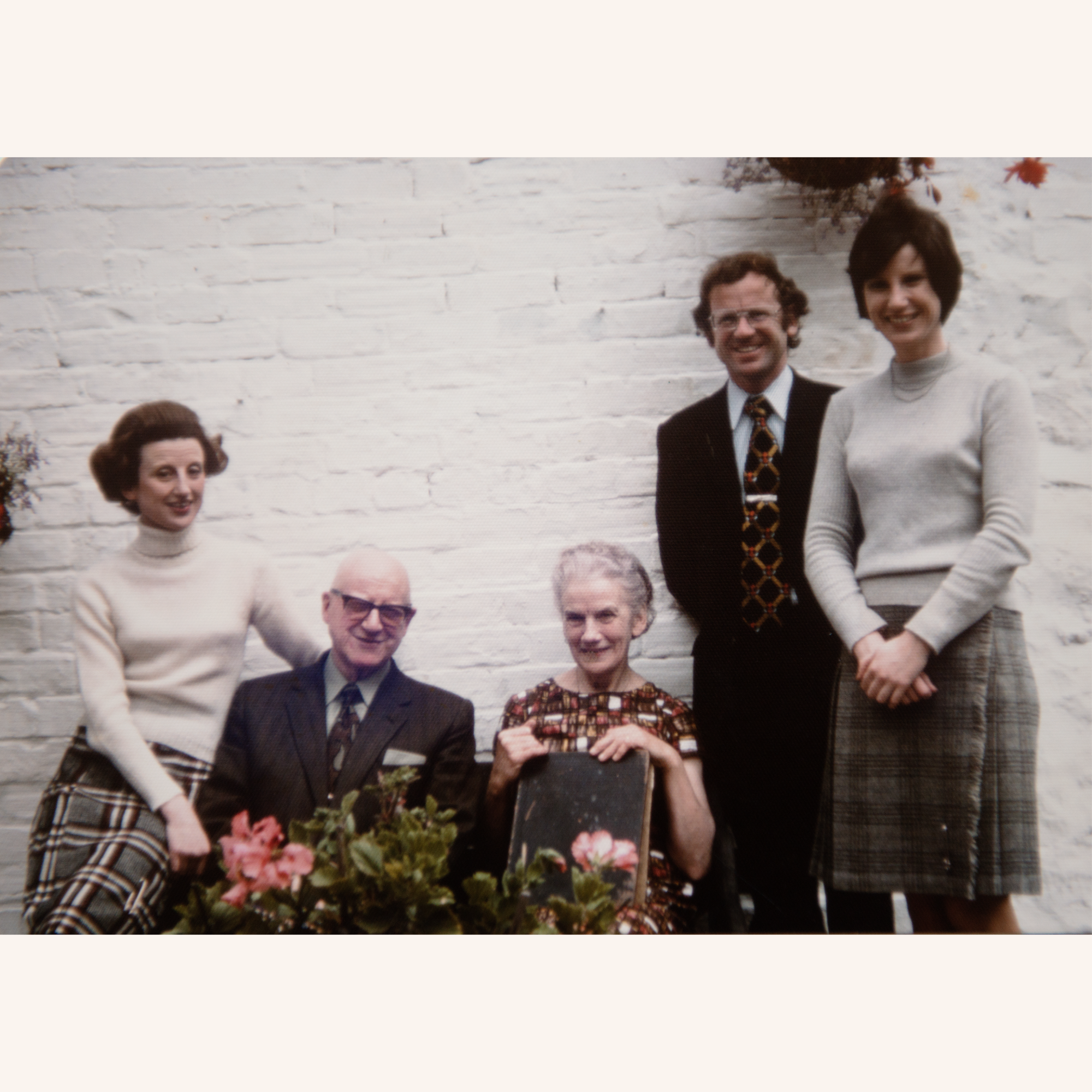 Five people posing together in front of a white brick wall, with two women and one man standing behind an older man and woman who are seated. Some flowers are in the foreground.