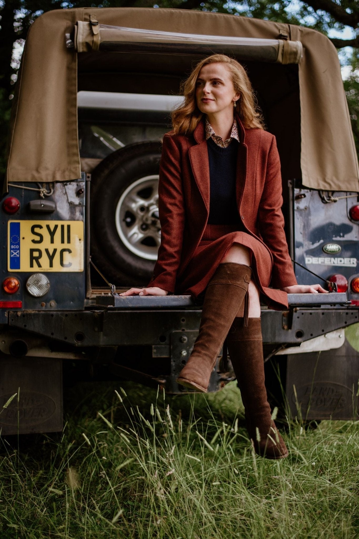 A woman with long, light brown hair sits on the tailgate of a Land Rover Defender, wearing the Campbells of Beauly House Jacket, surrounded by grass and trees.