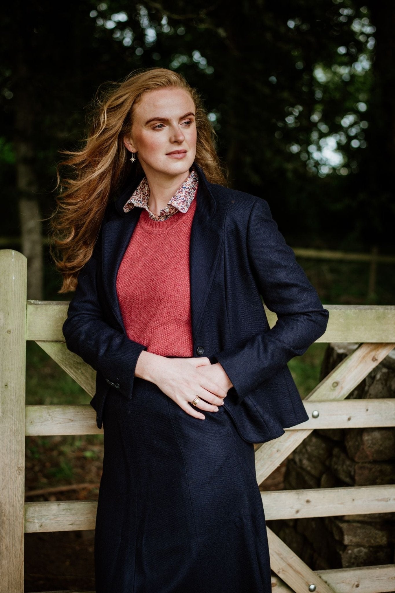 A woman with long, wavy hair stands outdoors by a wooden gate, wearing the House Jacket by Campbells of Beauly over a red sweater and floral-collared shirt. Trees and a fence appear in the background.
