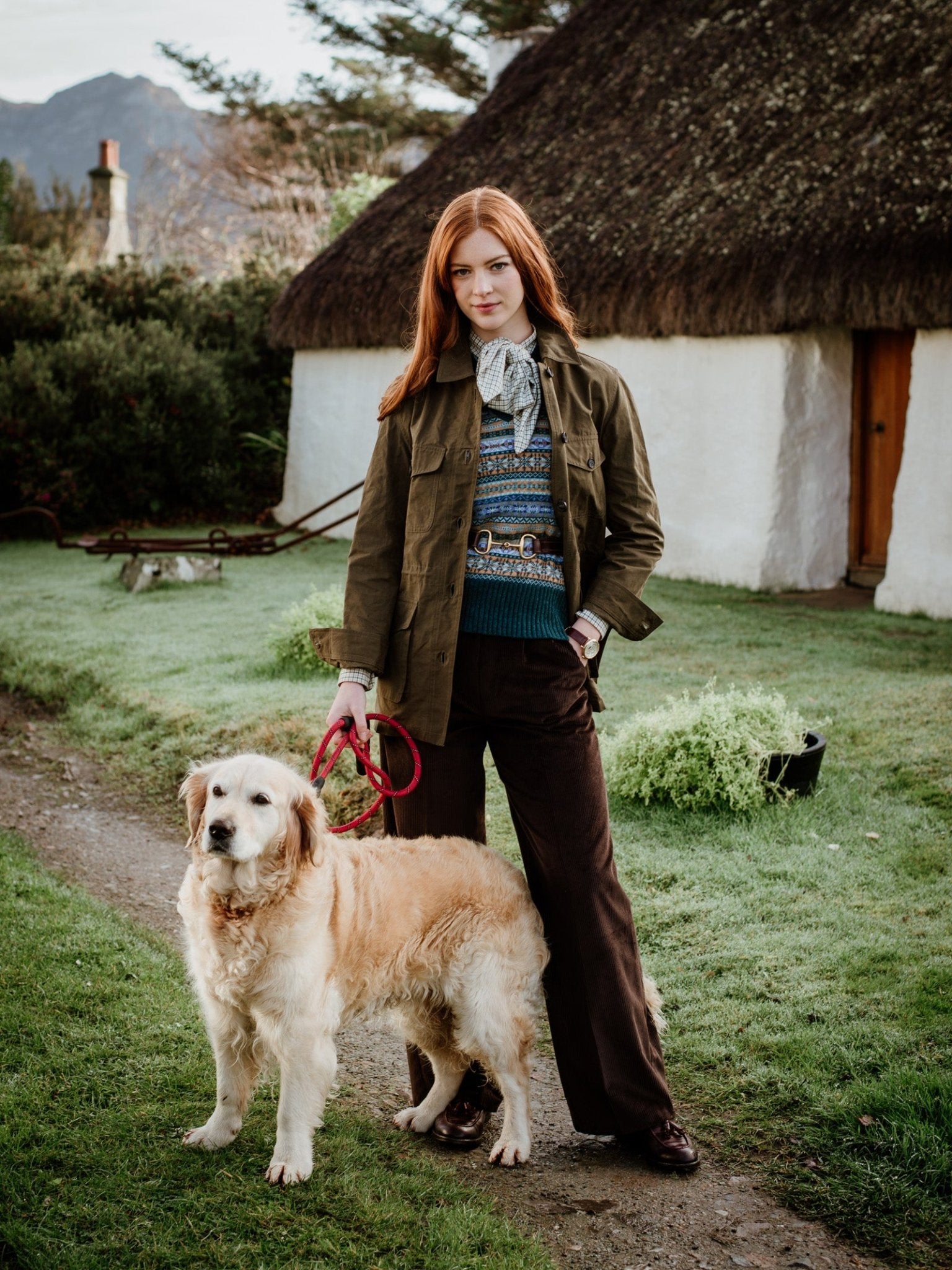 A woman with long red hair stands on a grassy path beside a golden retriever, wearing Campbells of Beauly Aigas Khaki Field Jacket, a patterned sweater, and brown pants. A white thatched cottage is visible in the background.