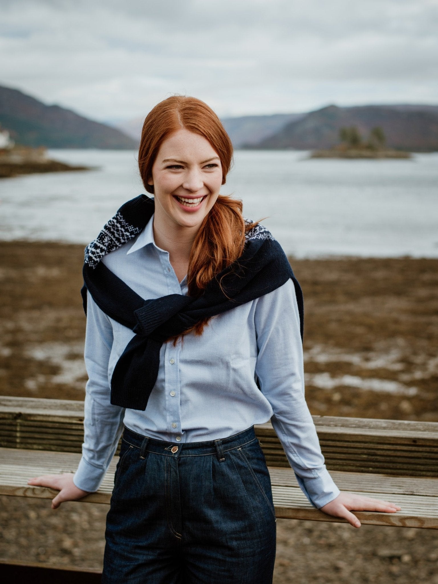 A smiling woman with long red hair wears a Campbells of Beauly Brushed Cotton Shirt in classic fit light blue and dark pants, standing outdoors by a wooden railing with a scenic lake and hills behind her.