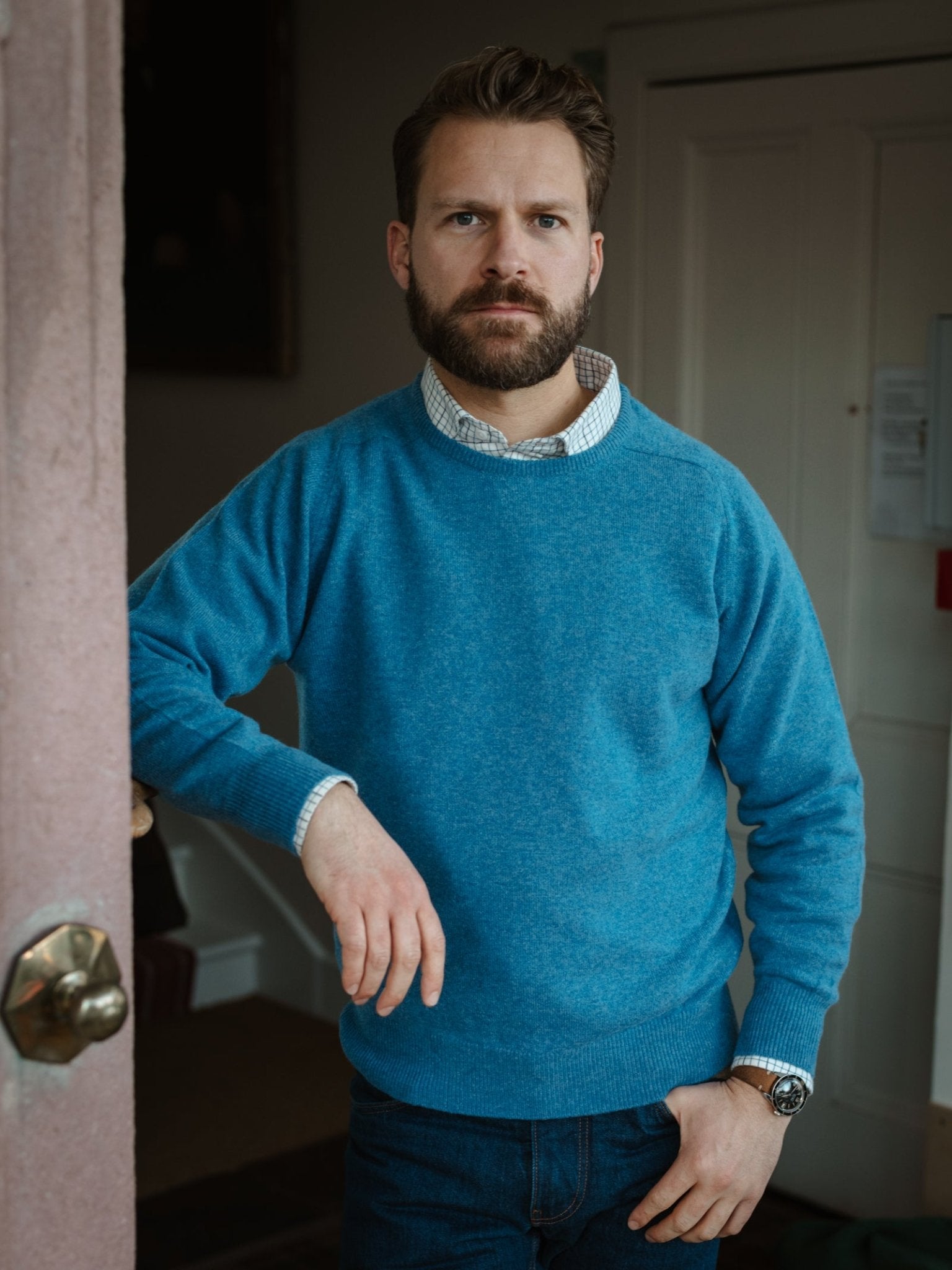 A bearded man stands indoors by a doorway, one hand on the frame and the other in his pocket, wearing Campbell's of Beauly's Lambswool Crew Neck Jumper over a collared shirt with jeans.