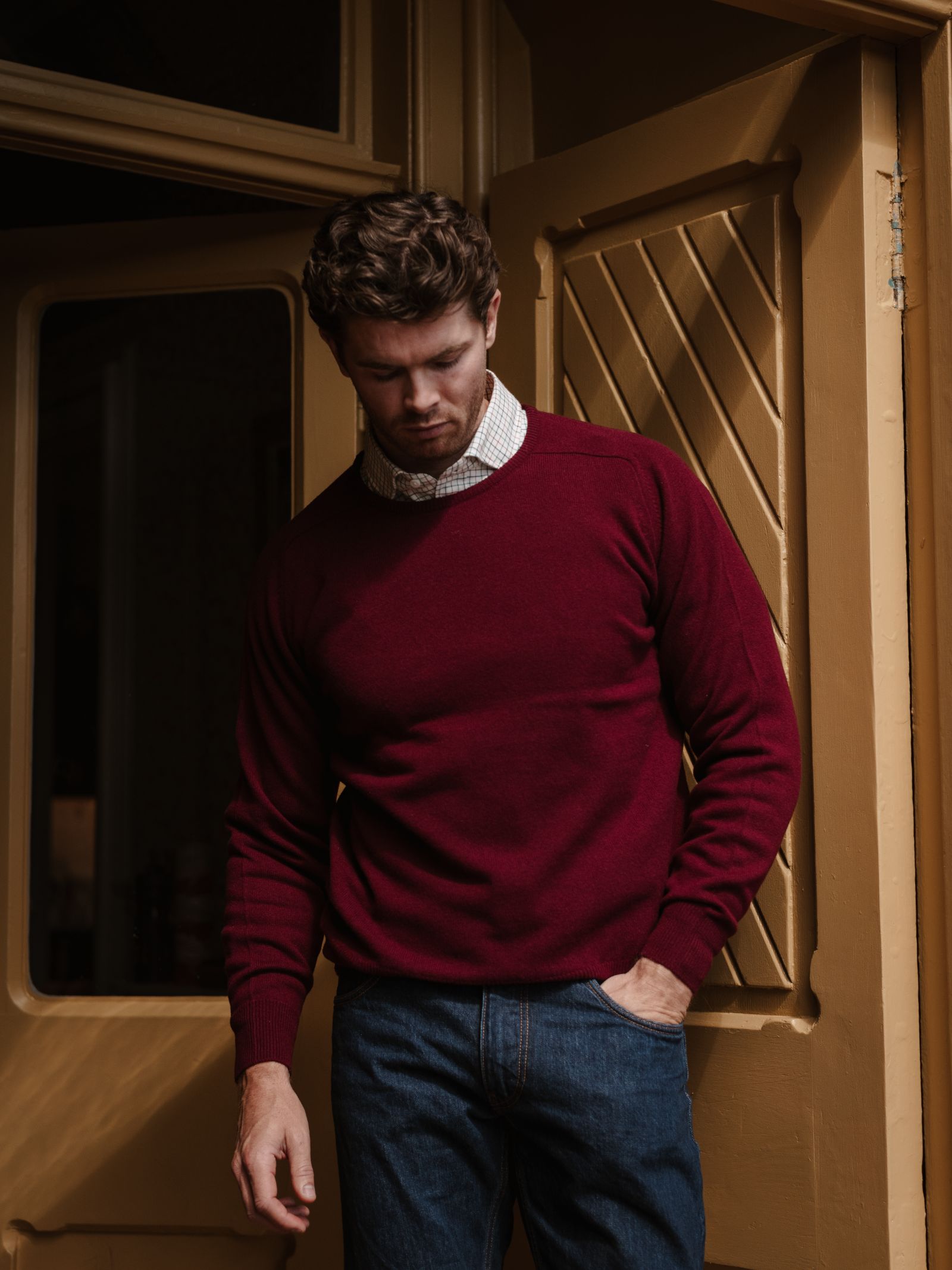 A man wearing a Campbell's of Beauly Lambswool Crew Neck Jumper over a collared shirt and blue jeans stands by a golden brown door, looking down with one hand in his pocket.