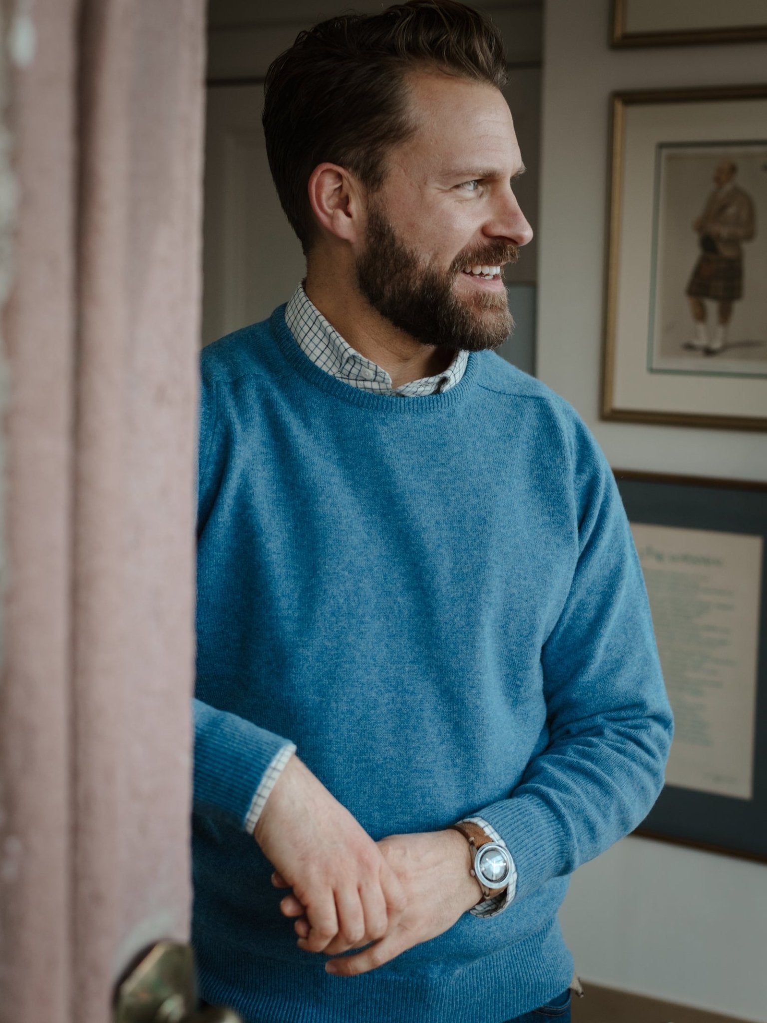 A bearded man stands indoors by a door, smiling and looking right. He's wearing a blue Campbell's of Beauly Lambswool Crew Neck Jumper over a plaid shirt and wristwatch; framed pictures are on the wall behind him.