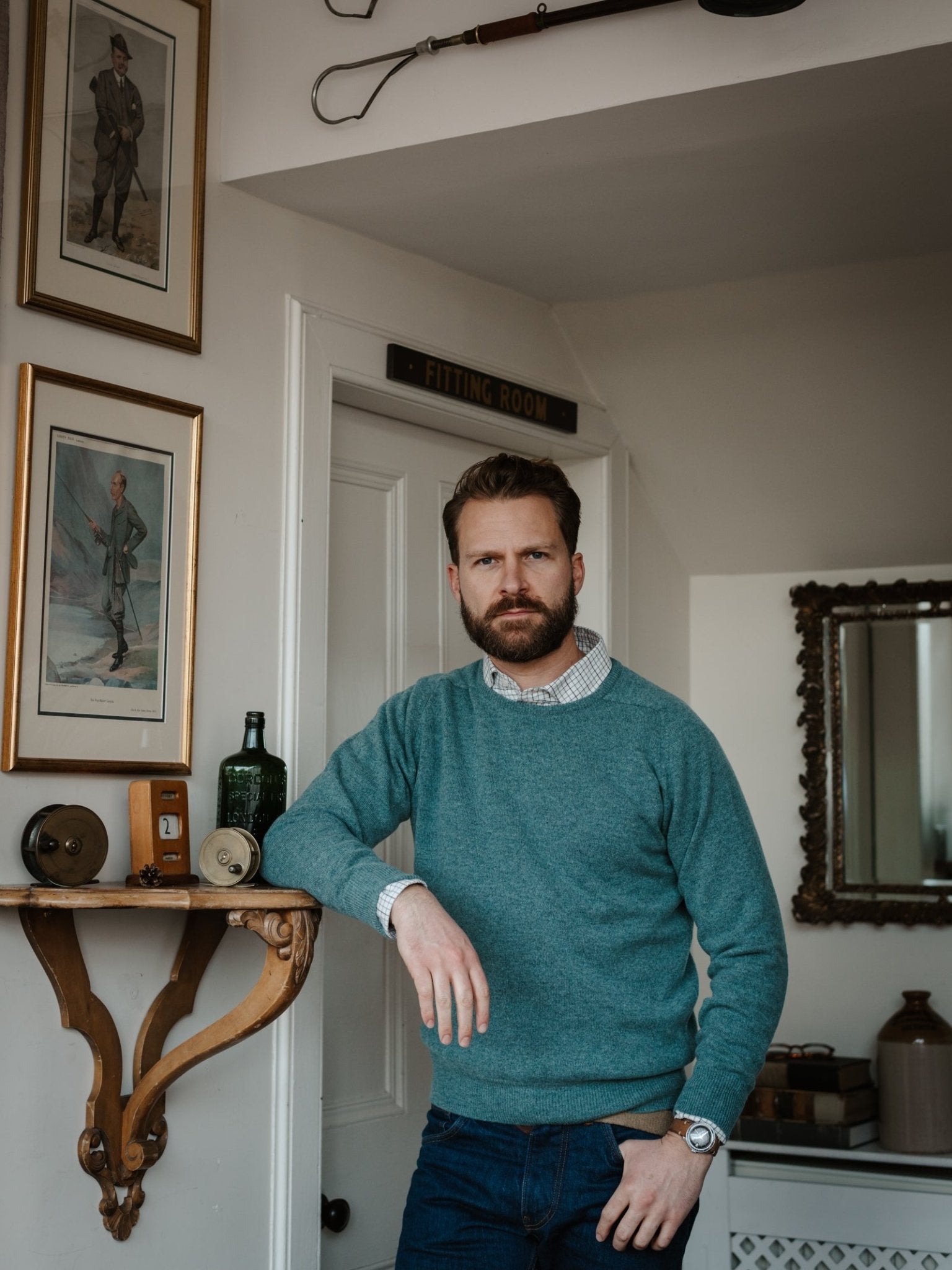 A bearded man in a teal Campbell's of Beauly Lambswool Crew Neck Jumper and jeans leans against a wooden shelf in a vintage room decorated with framed art, antiques, and a mirror.