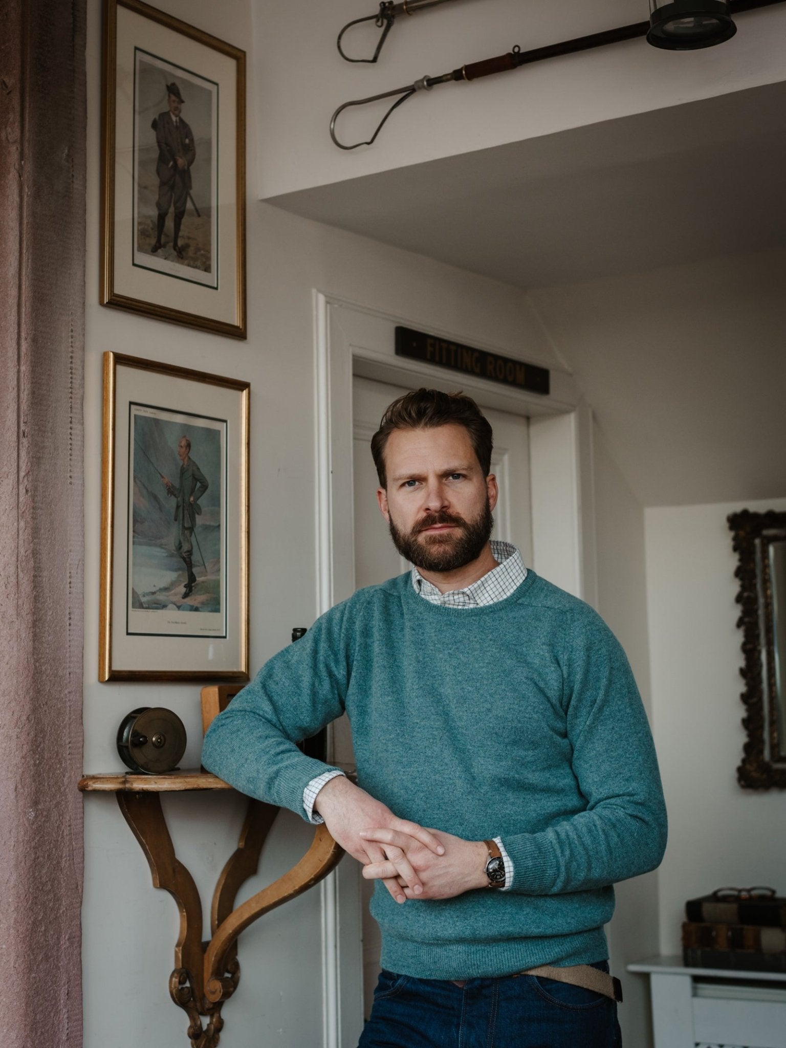 A bearded man stands indoors, leaning against a wooden shelf. He wears a Campbell's of Beauly Lambswool Crew Neck Jumper in teal over a checked shirt, with framed paintings, a mirror, and vintage objects displayed on the wall behind him.