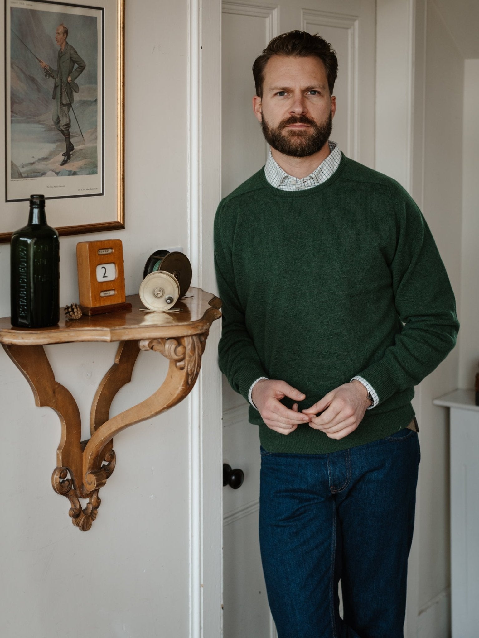 A bearded man in a Campbell's of Beauly Lambswool Crew Neck Jumper and blue jeans stands indoors next to a small wall shelf with bottles, a clock, and a cup, under a framed painting.