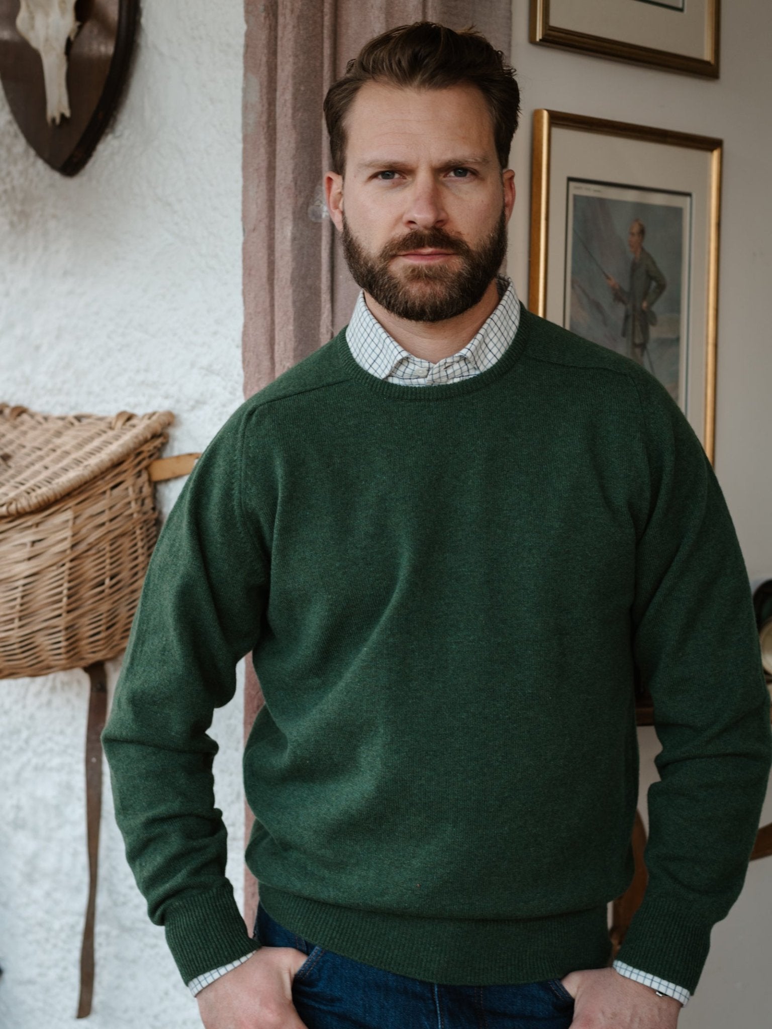 A bearded man in a Campbell's of Beauly Lambswool Crew Neck Jumper over a checked shirt stands indoors by a white wall with framed pictures and a woven basket.