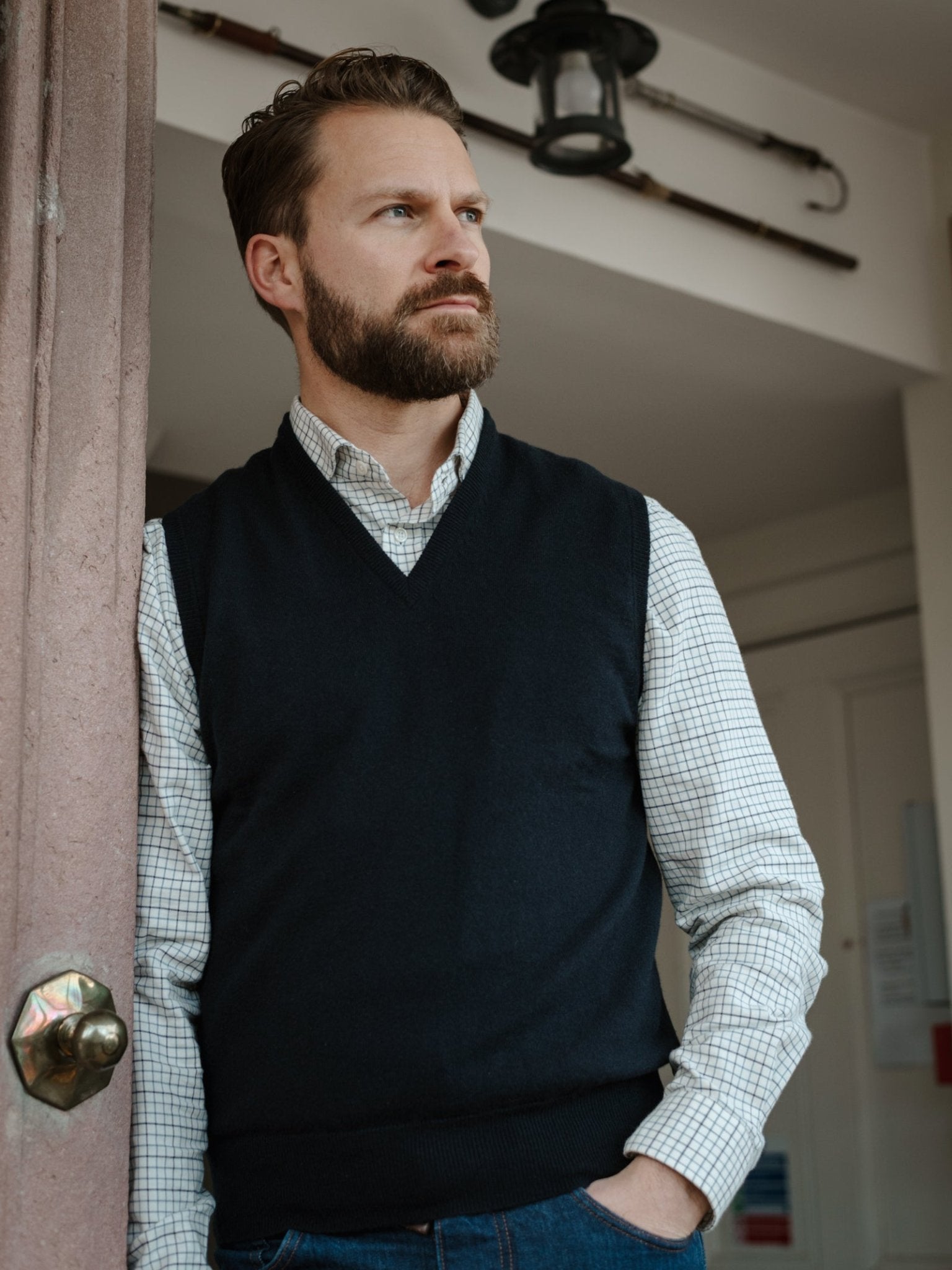 A man with a beard stands indoors by a doorway, wearing a checkered shirt and the Campbell's of Beauly Auld Stock Lambswool V-Neck Slipover, one hand in his pocket, gazing thoughtfully into the distance.