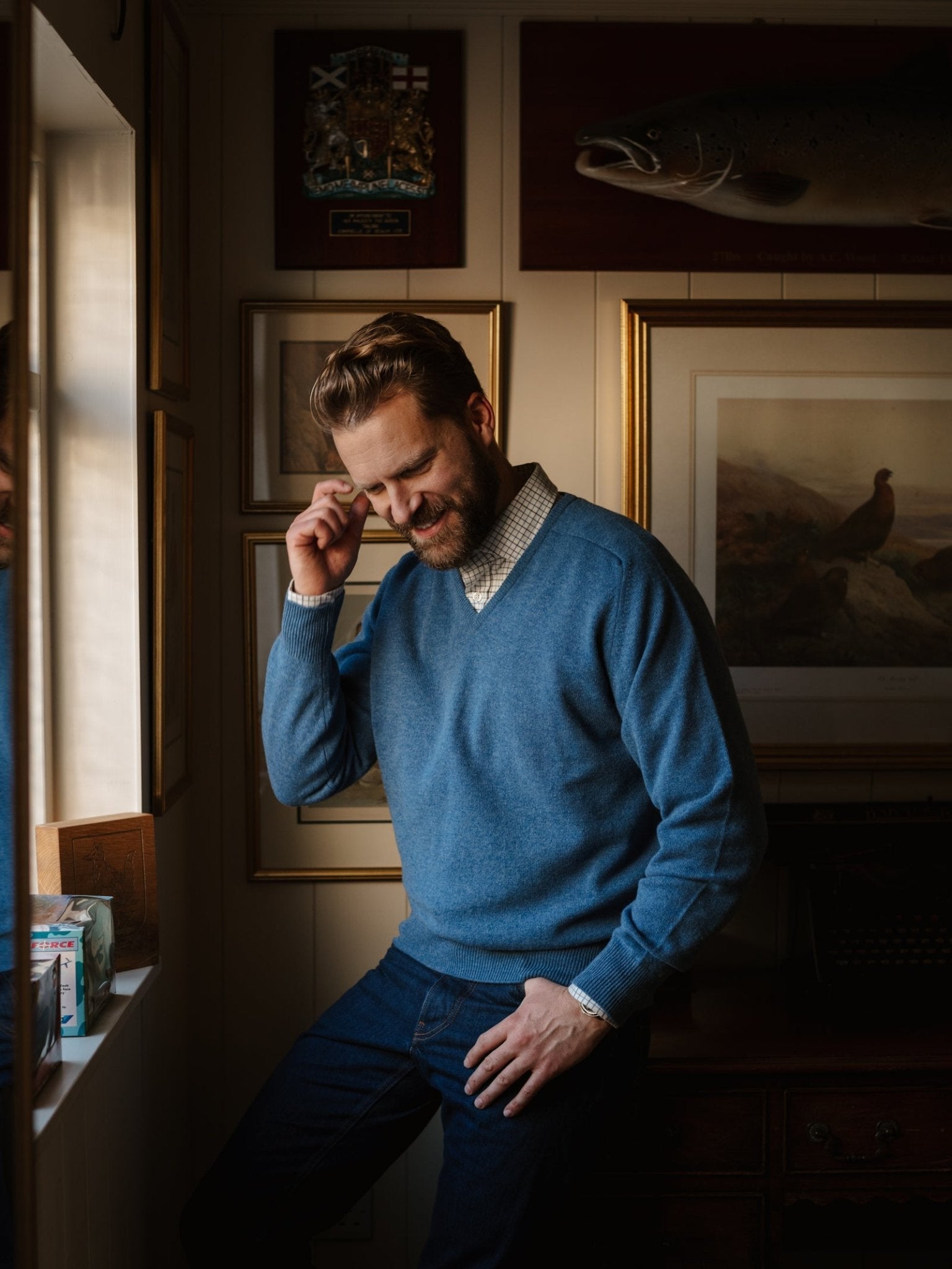 A bearded man in a Campbell's of Beauly Lambswool V-Neck Jumper and jeans stands by a window, smiling and touching his temple, in a cozy room with books, framed pictures, and a mounted fish on the wall.