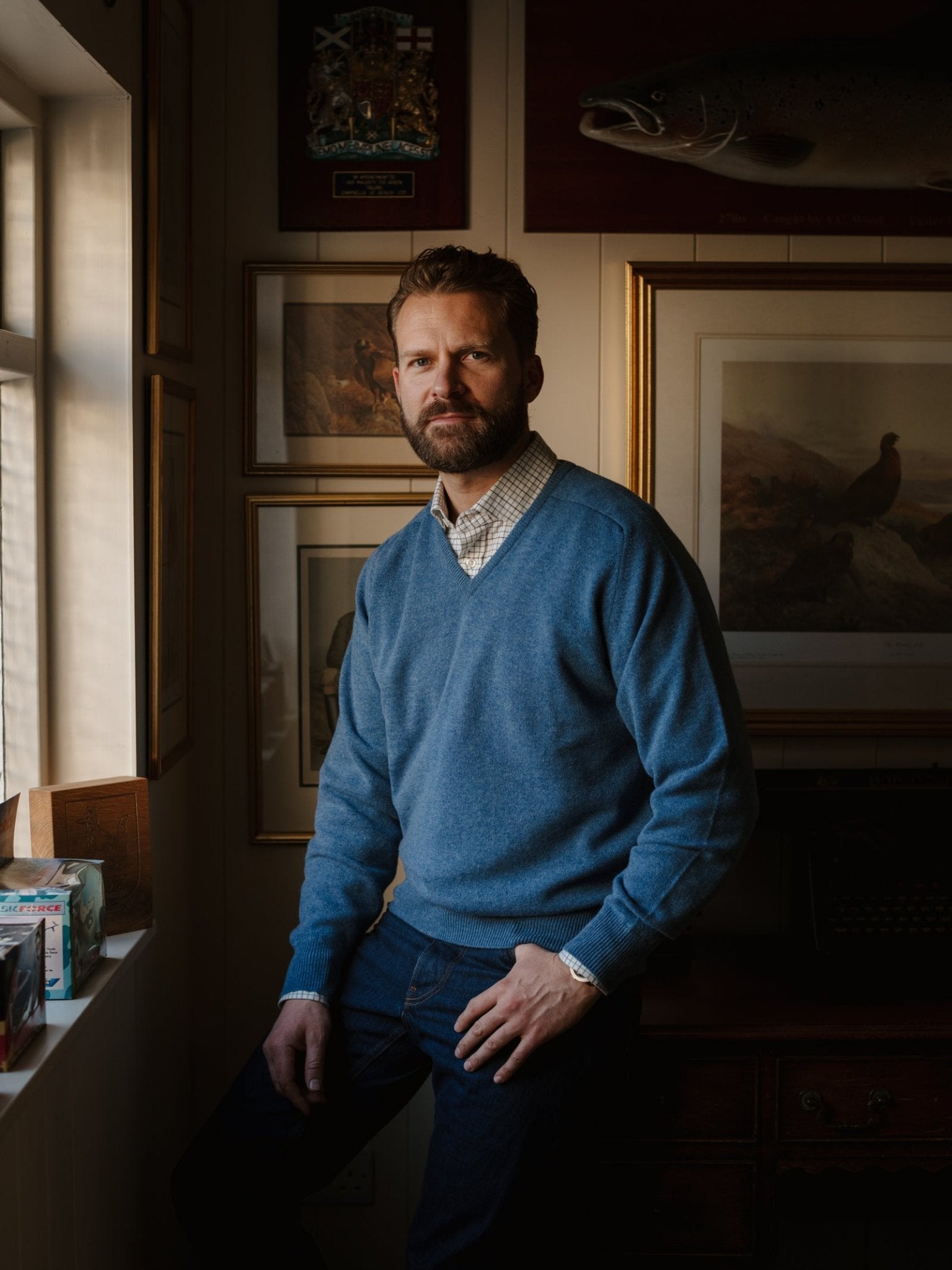 A bearded man wearing a Campbell's of Beauly Lambswool V-Neck Jumper and jeans sits on a windowsill in a warmly lit room decorated with framed art, photos, and a mounted fish on the wall.