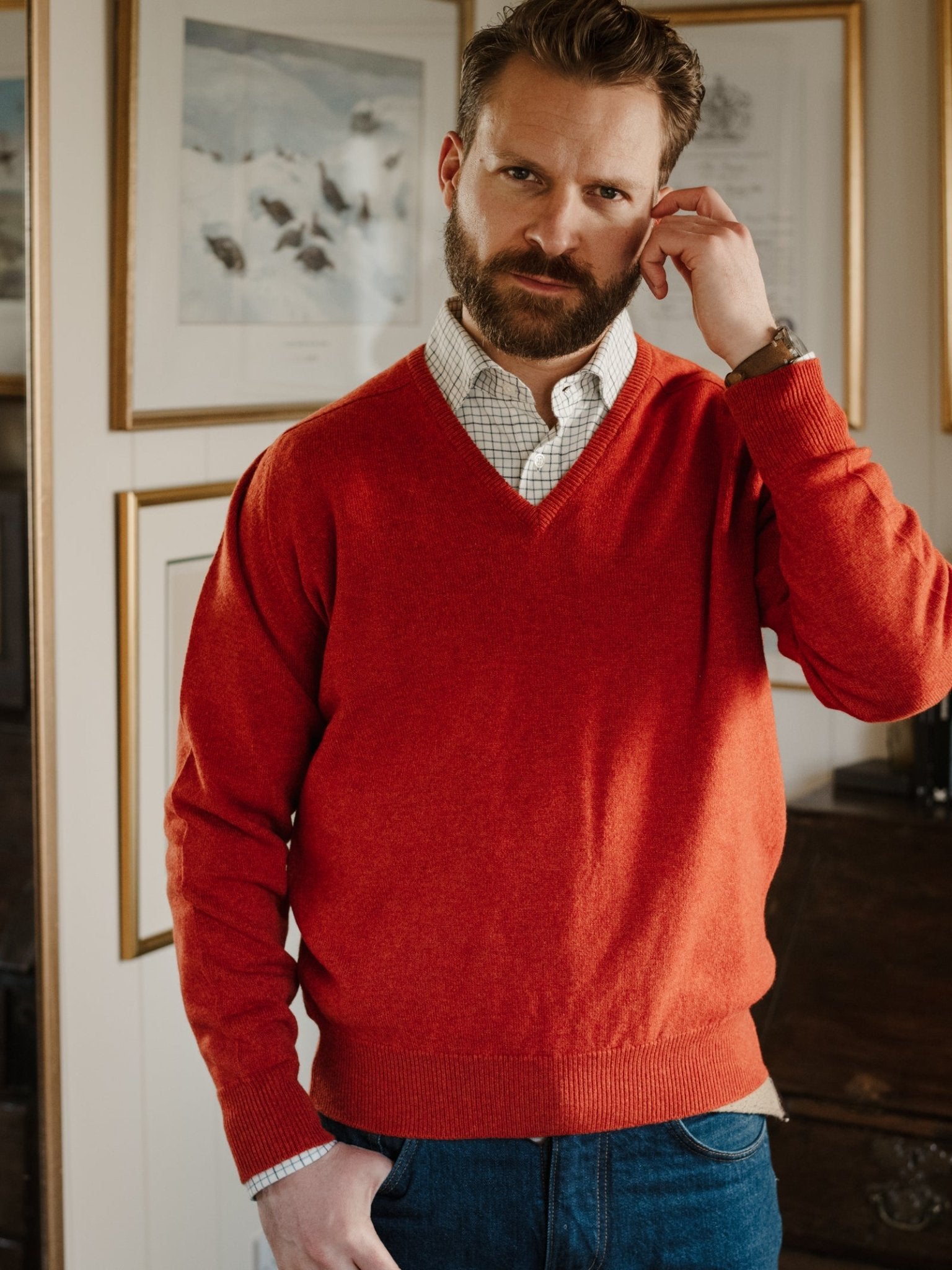 A bearded man indoors touches his ear, wearing Campbell's of Beauly Lambswool V-Neck Jumper in red over a checked shirt and blue jeans, with framed artwork and a wooden cabinet in the background.