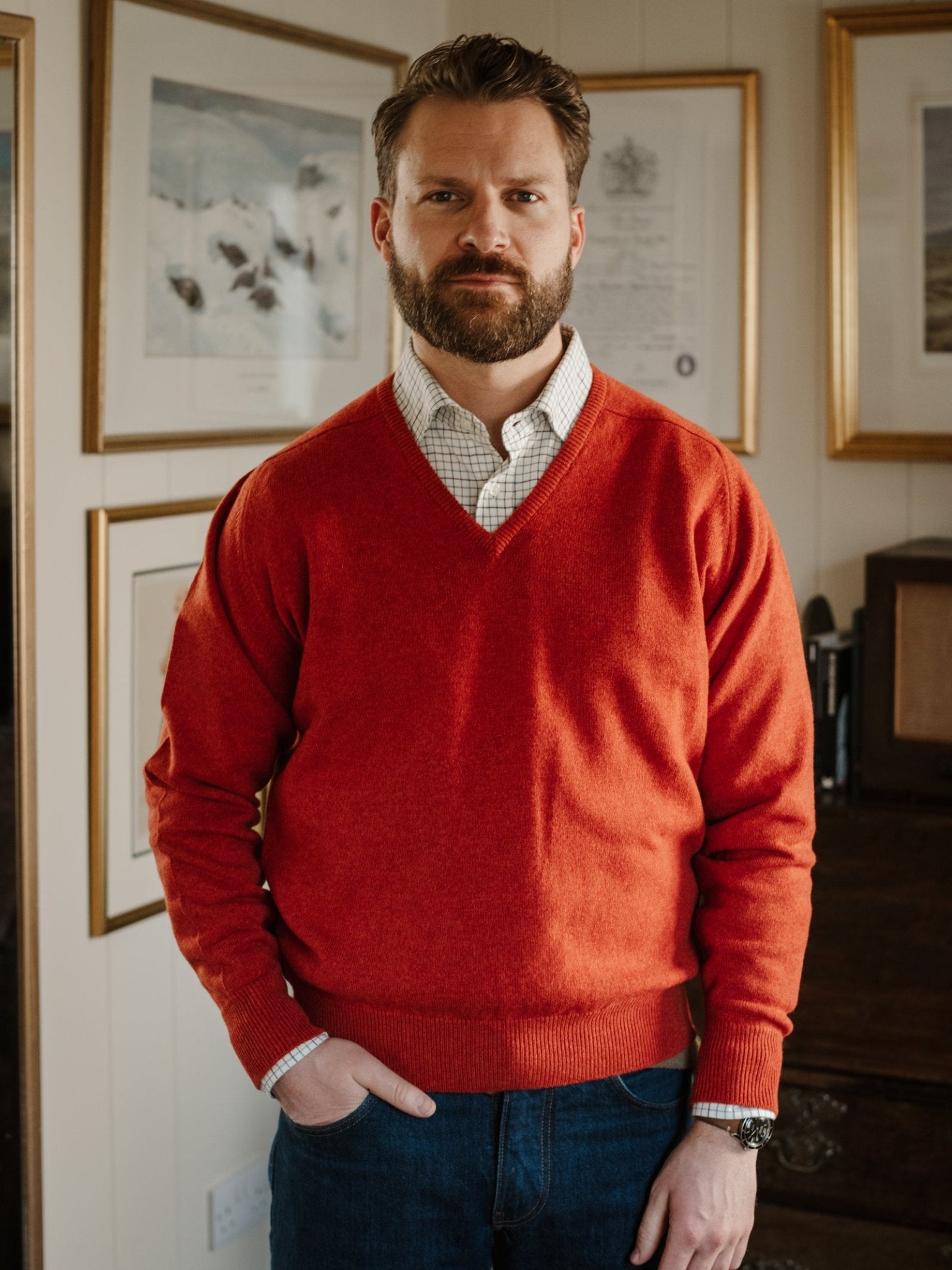 A bearded man stands indoors, one hand in his pocket, wearing the Campbell's of Beauly Lambswool V-Neck Jumper in red over a checked shirt and blue jeans, with framed pictures displayed on the wall behind him.