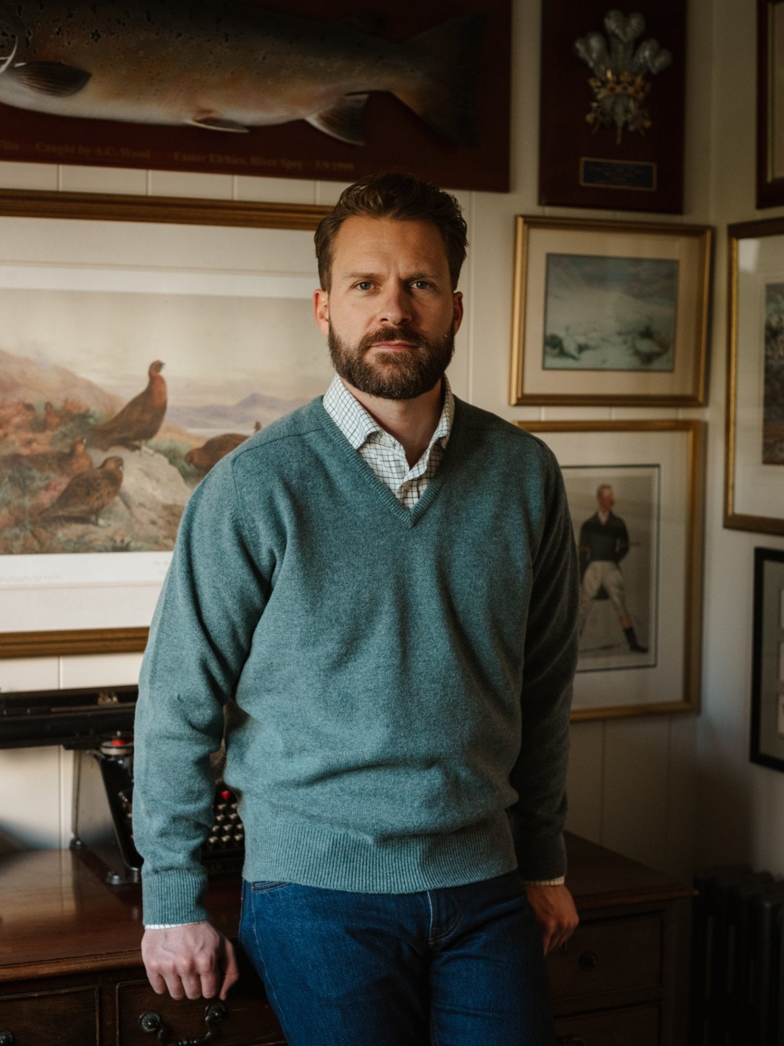 A bearded man wearing Campbell's of Beauly Lambswool V-Neck Jumper and jeans leans against a desk in a room decorated with framed art and photographs.