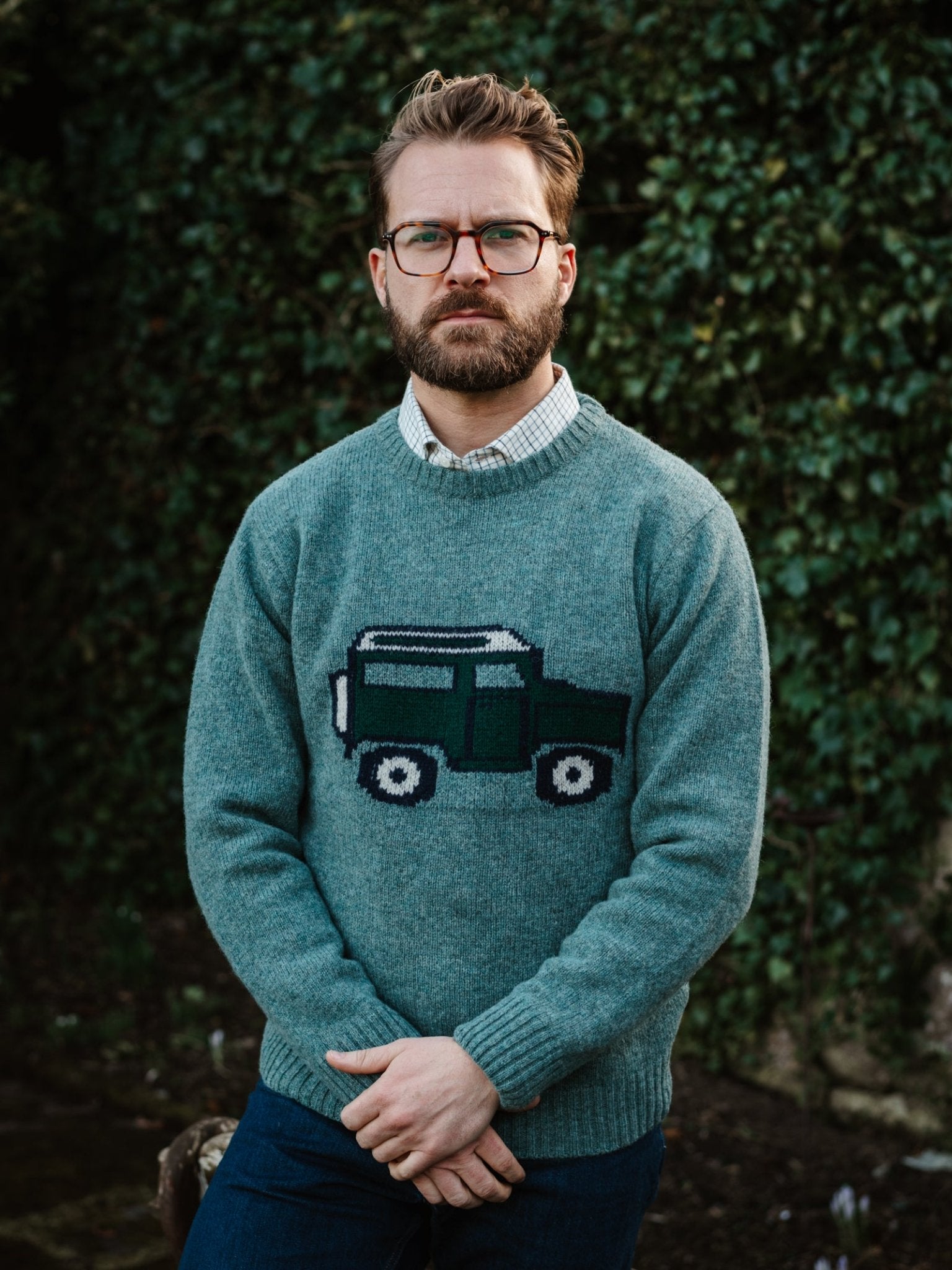 A man with glasses and a beard stands outdoors before lush foliage, wearing a light blue Unisex Land Rover Defender Jumper by Campbell's of Beauly—classic knitwear featuring a knitted vehicle design—over a collared shirt.