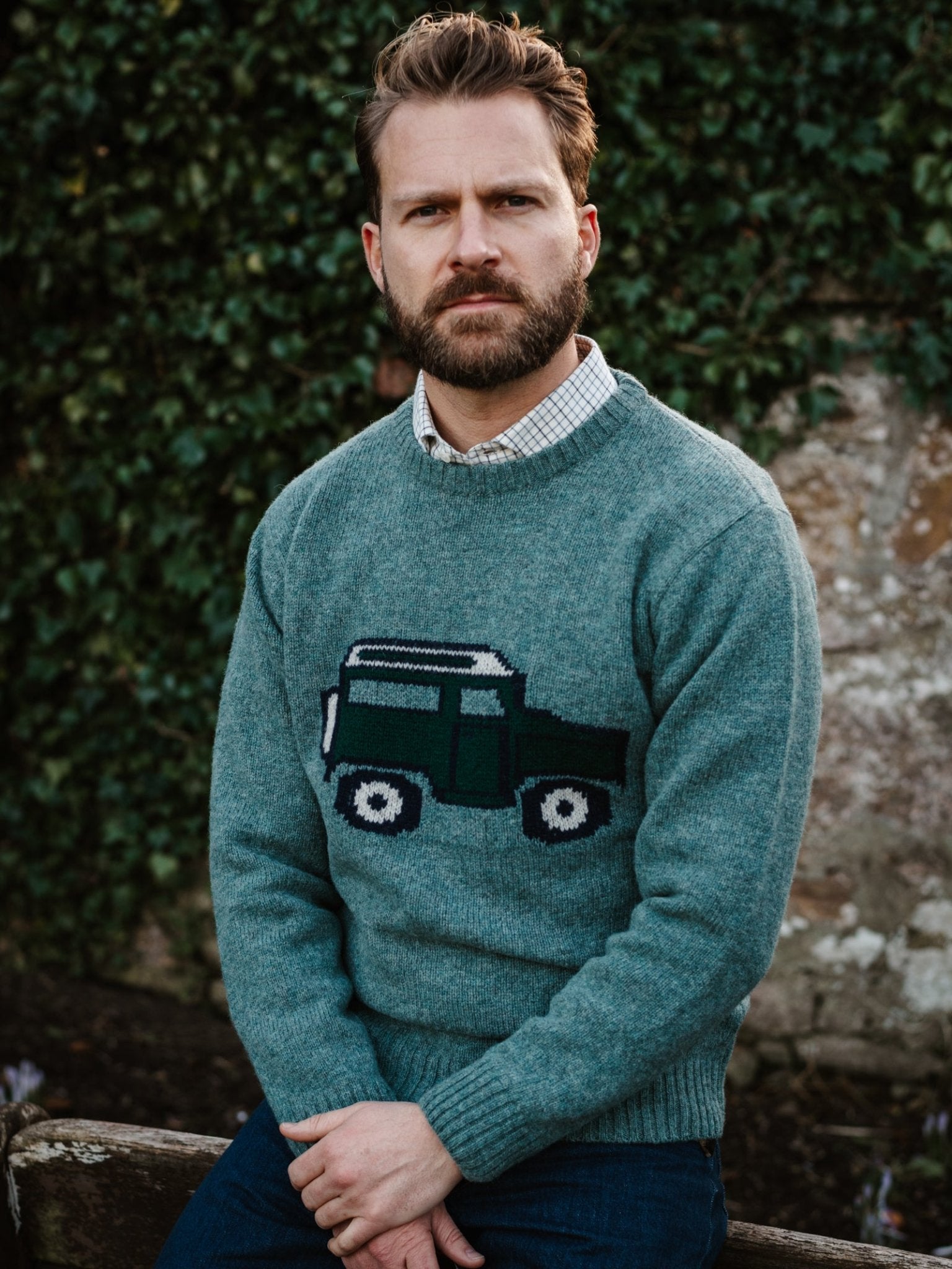 A man with short hair and a beard sits on a bench outdoors, wearing a green Unisex Land Rover Defender Jumper by Campbell's of Beauly. He looks serious, with green ivy and a stone wall in the background.