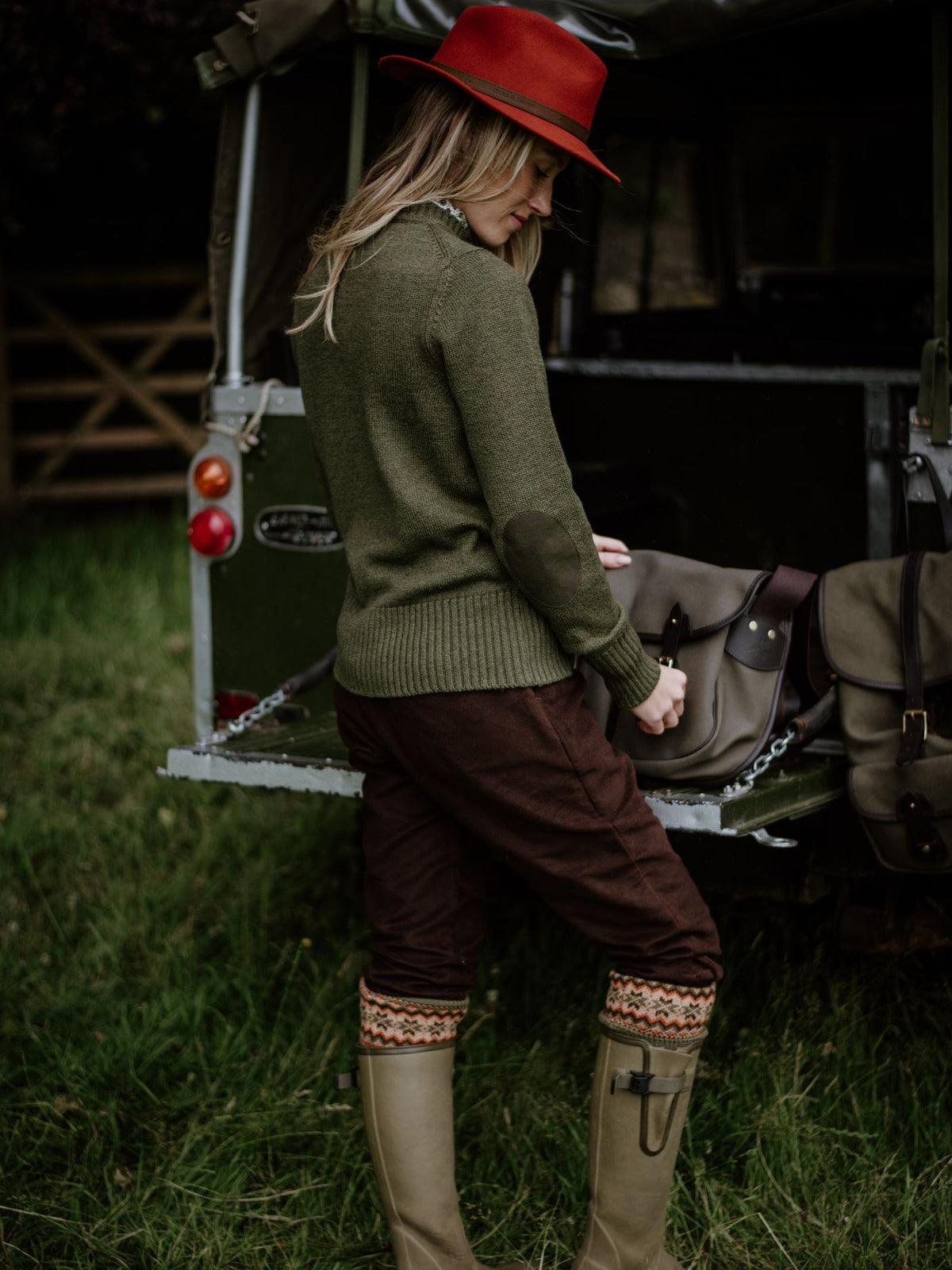 Dressed in Campbells of Beauly Moleskin Plus Twos, a person stands beside an open vehicle trunk outdoors, adjusting a bag on the grass—styled for country pursuits with olive green knitwear, patterned boots, and a red hat.