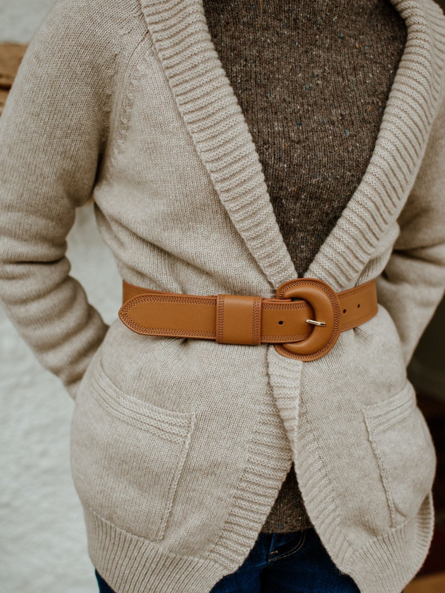 A person styles a beige knit cardigan with front pockets over a brown sweater, cinched with the Campbell’s of Beauly Caramel Leather Belt handmade in Italy, and paired with blue jeans.