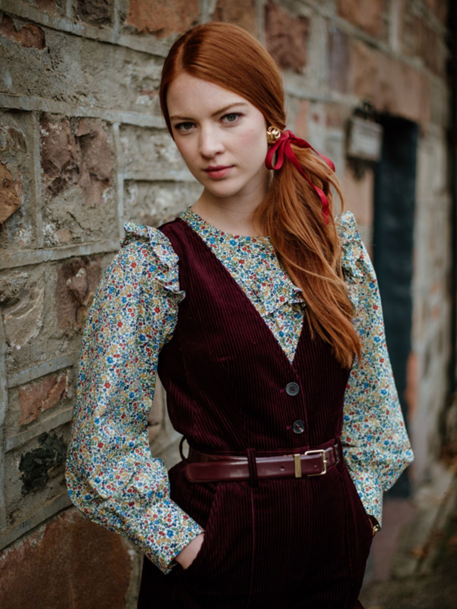 A young woman with long red hair tied with a red ribbon stands against a stone wall, wearing Campbells of Beauly Liberty Print Peplum Shirt with ruffled sleeves, paired with a burgundy corduroy vest and pants. She looks directly at the camera.