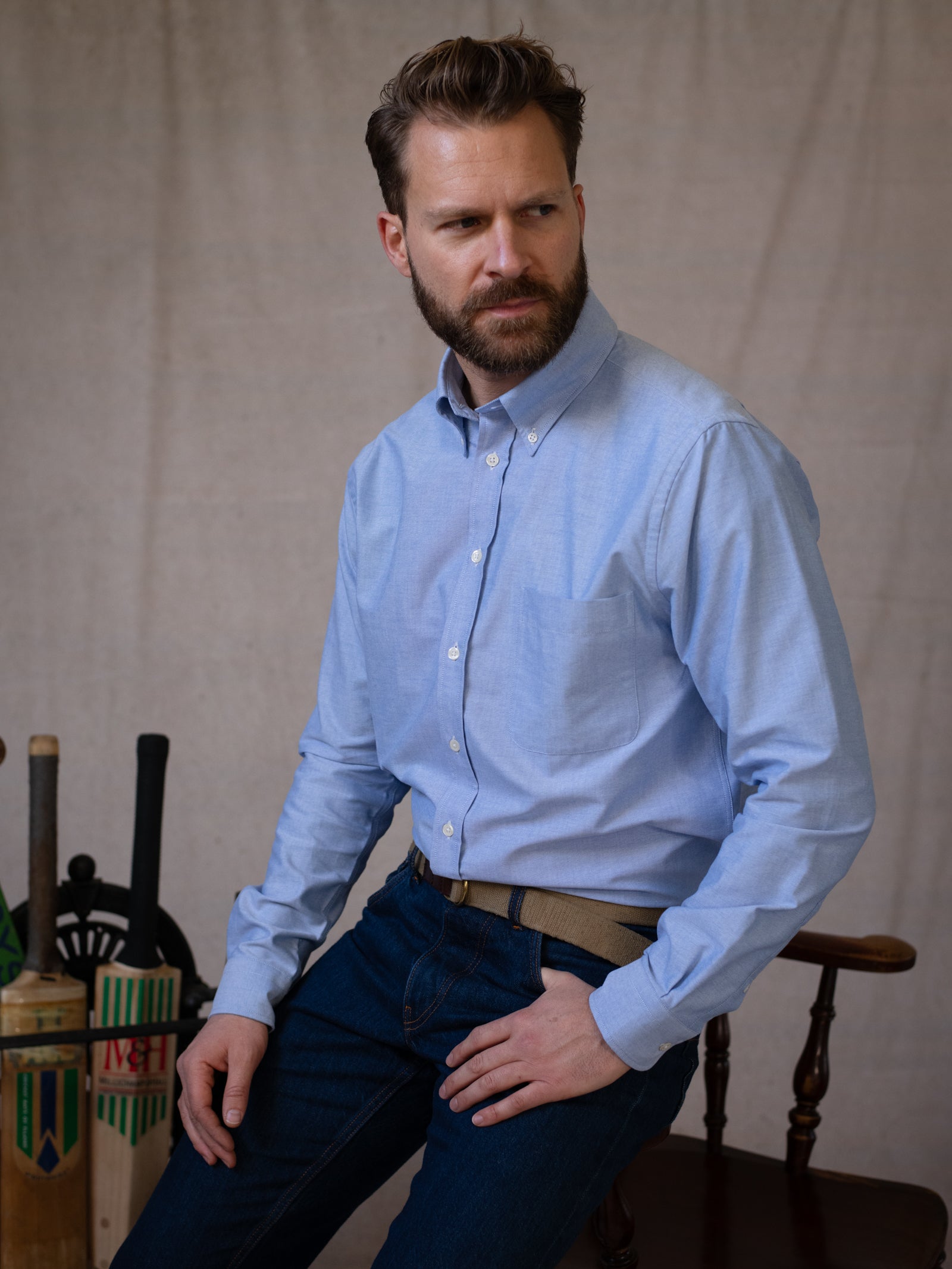 A bearded man with short hair wears a light blue Campbell’s of Beauly Oxford Button Down Shirt and jeans while seated on a wooden chair. Cricket bats and stumps are beside him against a plain backdrop.