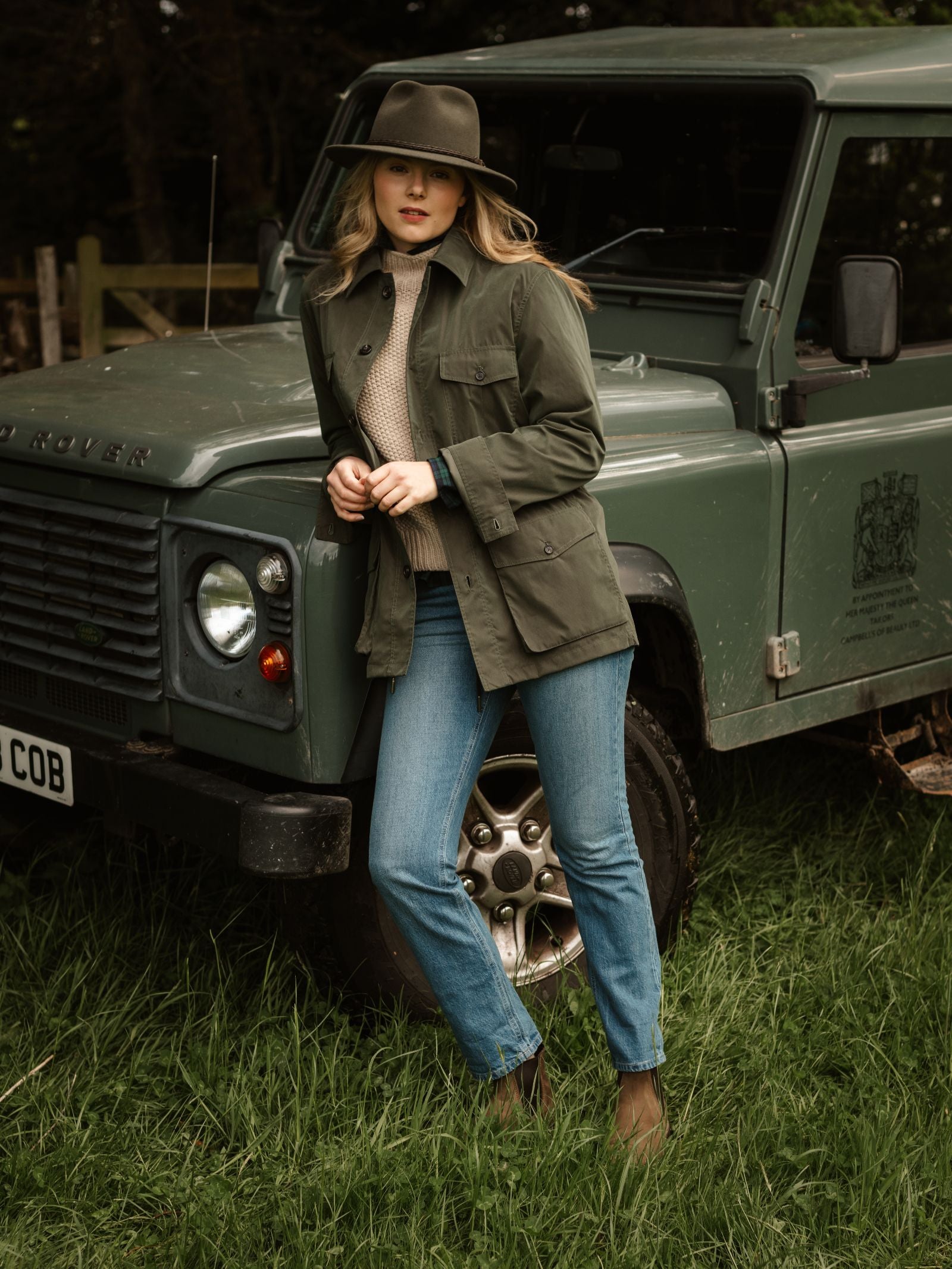 A woman in a Campbell's of Beauly Lightweight Aigas Field Jacket, hat, and blue jeans leans against a green Land Rover Defender on grass, with trees and a wooden fence behind her.