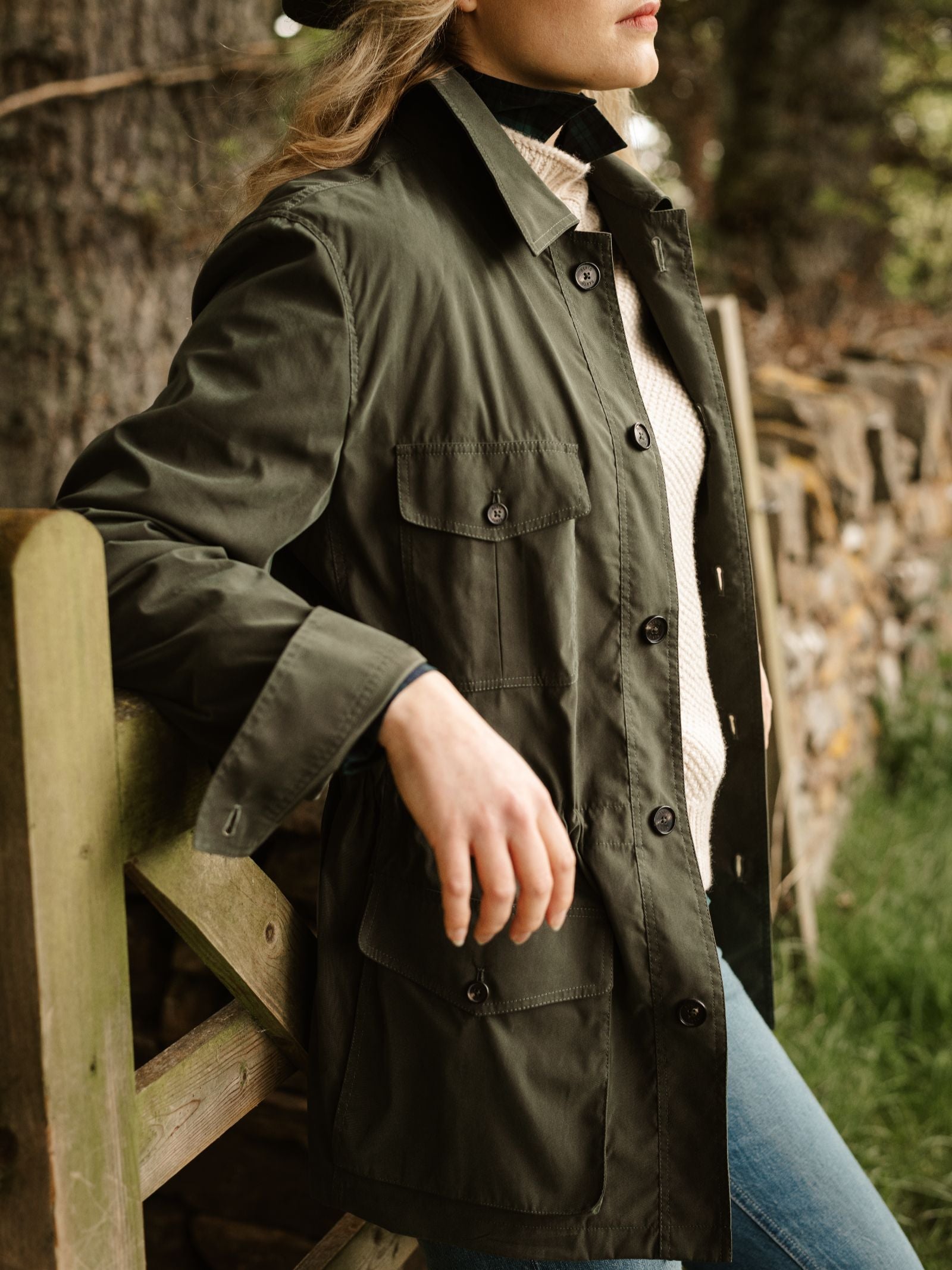 A person in a green Lightweight Aigas Field Jacket by Campbell's of Beauly and blue jeans leans on a wooden fence outdoors, with greenery and a stone wall behind them. Their face is partially out of frame.
