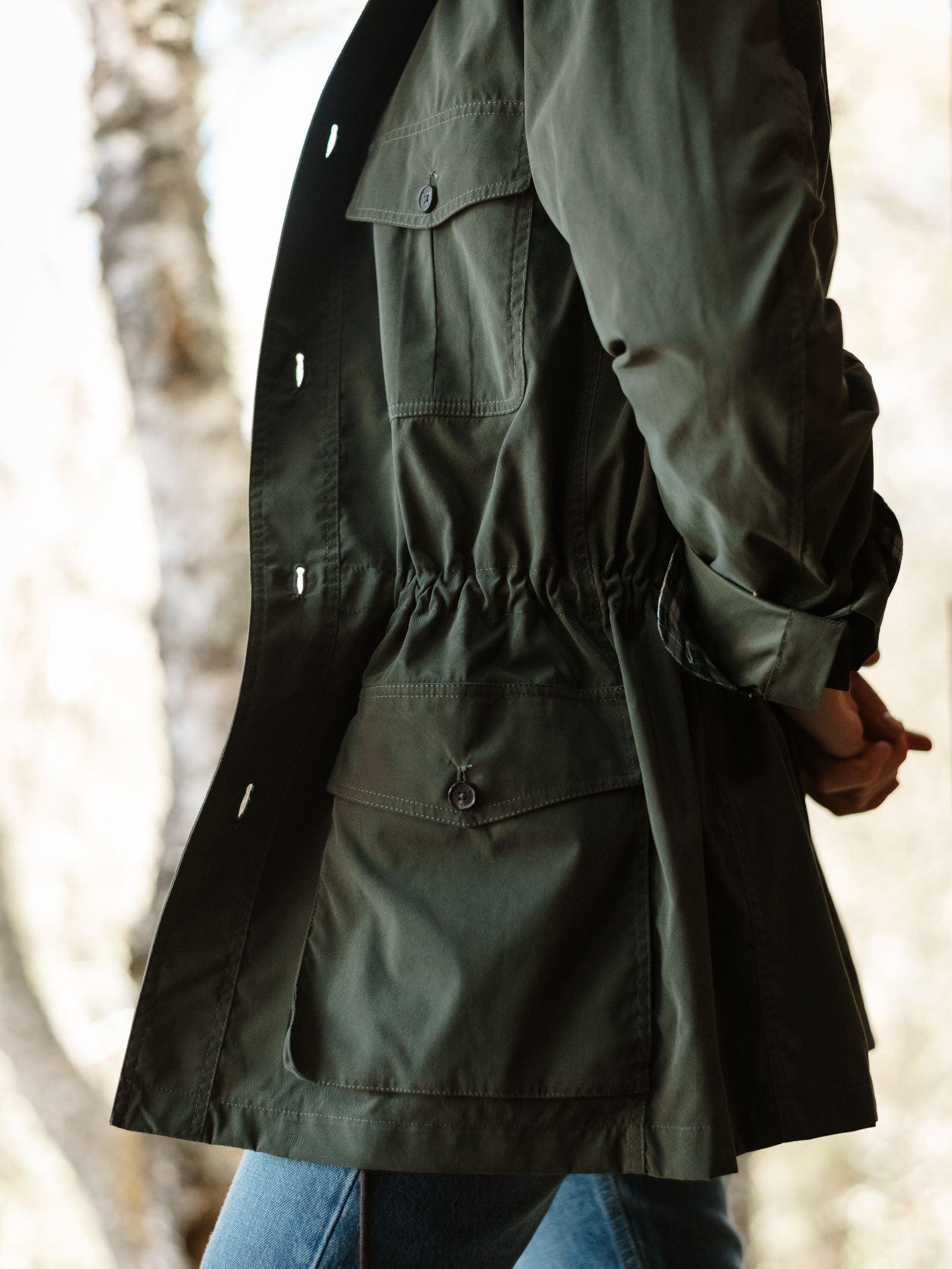 A person wearing a Campbell's of Beauly Lightweight Aigas Field Jacket in dark green and blue jeans stands outdoors, with blurred trees in the background.