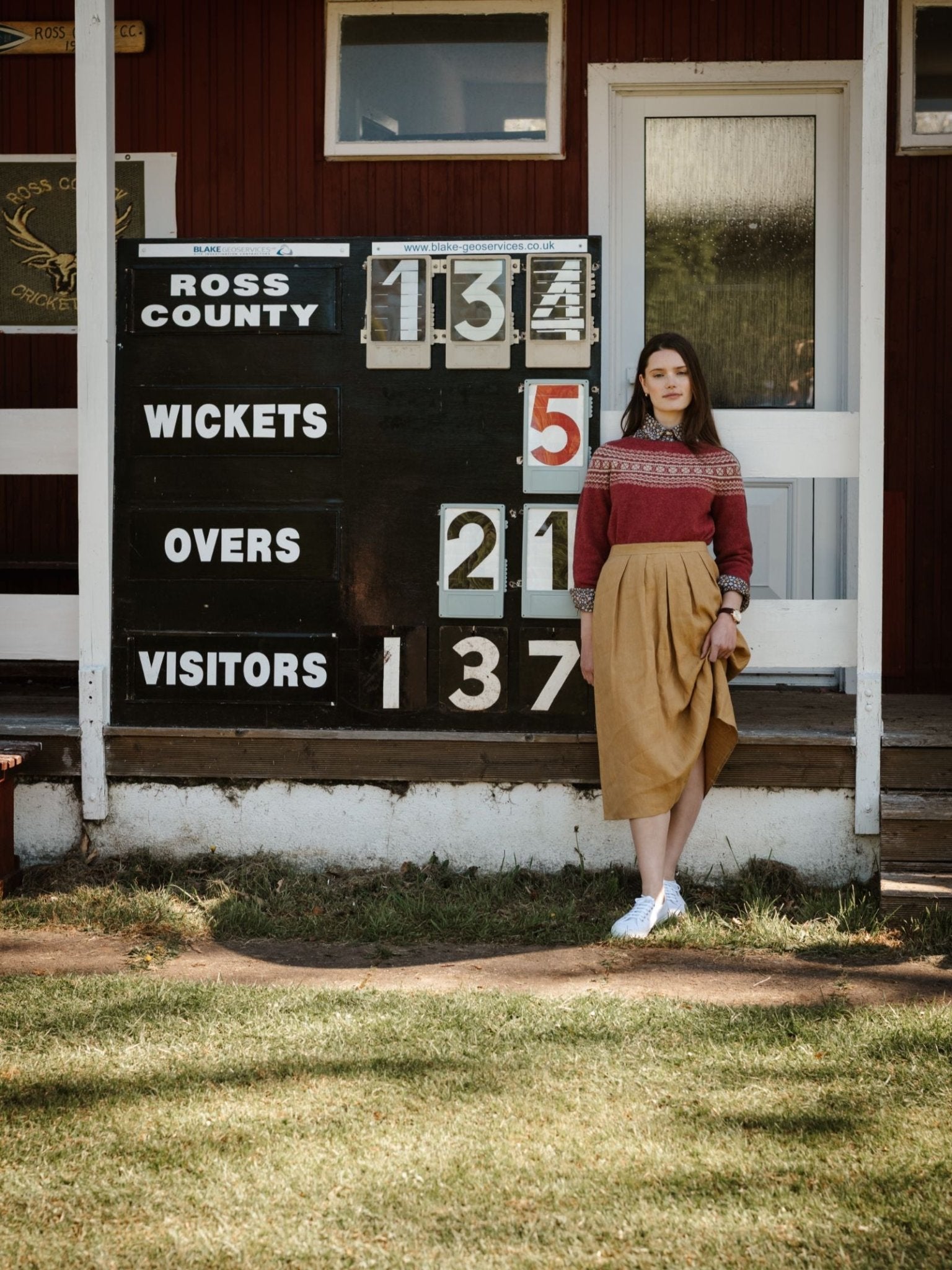 A woman wearing a Campbell's of Beauly Linen Skirt and red sweater stands by a vintage cricket scoreboard (Ross County: 134/5 in 21 overs, Visitors: 137) outdoors, with a wooden building behind her.