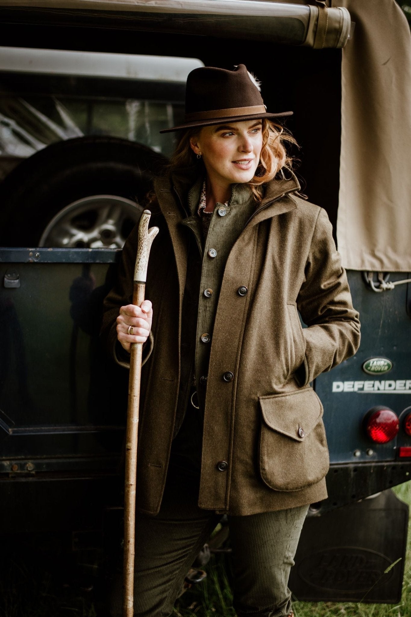 A woman in a classic style, wearing a brown hat and Campbell's of Beauly Loden Green Field Coat, stands by a Land Rover Defender with a wooden walking stick, smiling and ready for adventure.