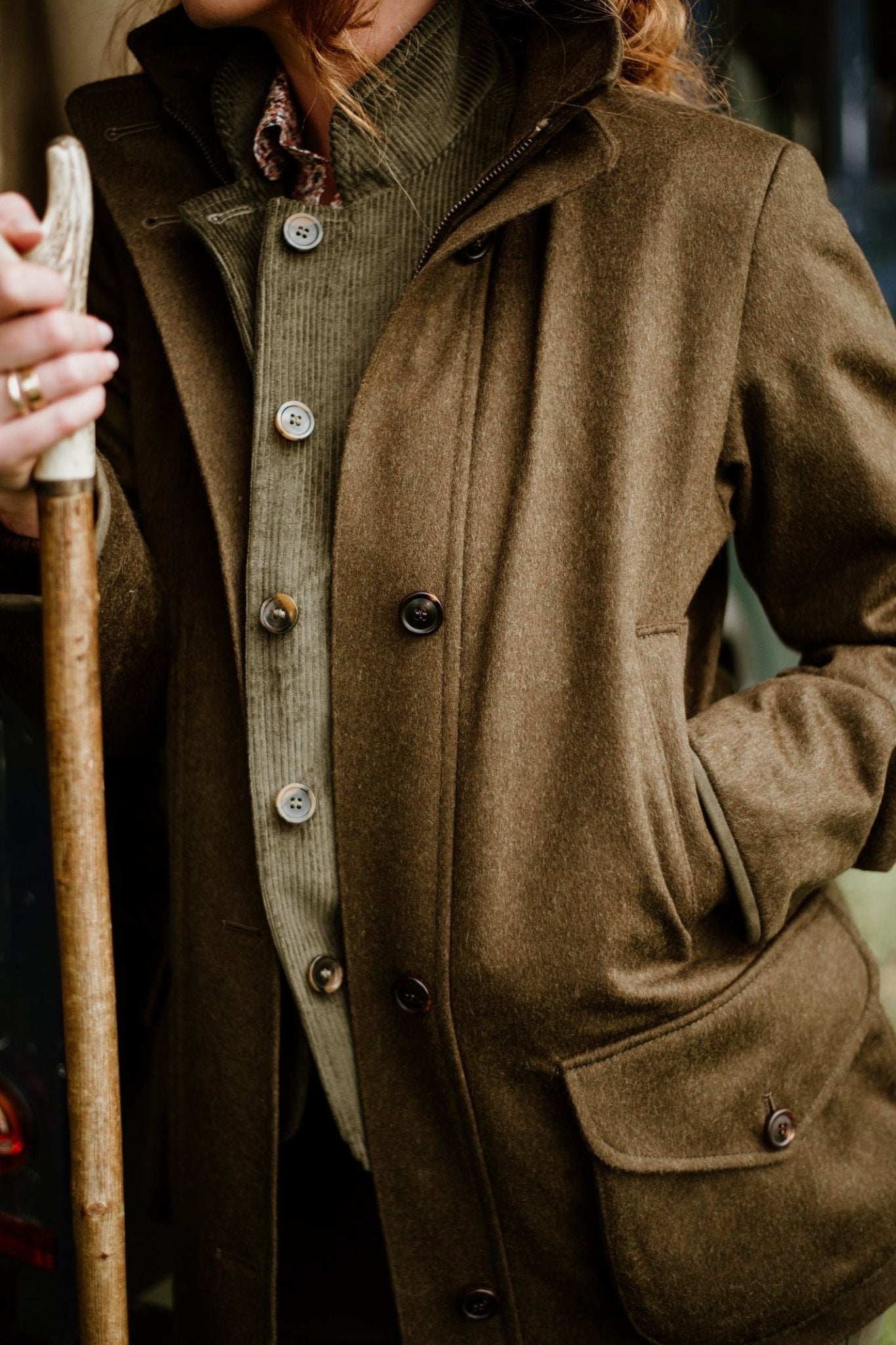Wearing Campbells of Beauly Loden Green Field Coat and a buttoned cardigan, a person stands with one hand in their pocket and the other on a wooden stick. Their face is partially out of frame.
