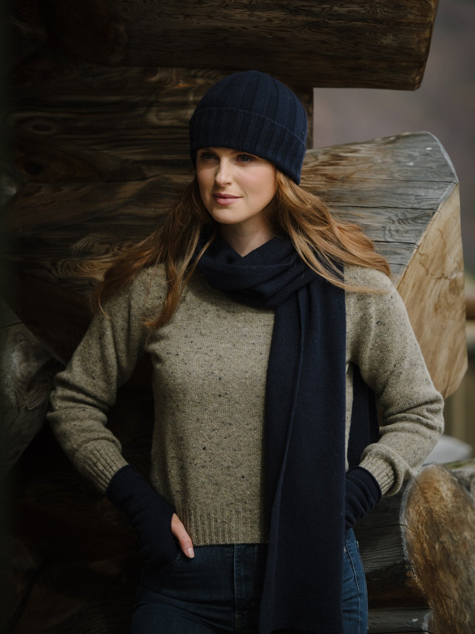 A woman wearing Campbells of Beauly Cashmere & Merino Fingerless Mittens, a navy knit hat, and a merino wool scarf with a beige sweater and jeans stands before large wooden beams, gazing slightly to the side.