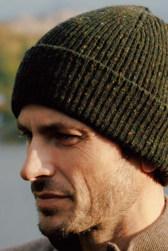 A man wearing the Campbells of Beauly Donegal Lambswool Hat in green and a brown sweater gazes thoughtfully into the distance, set against a softly blurred outdoor backdrop.