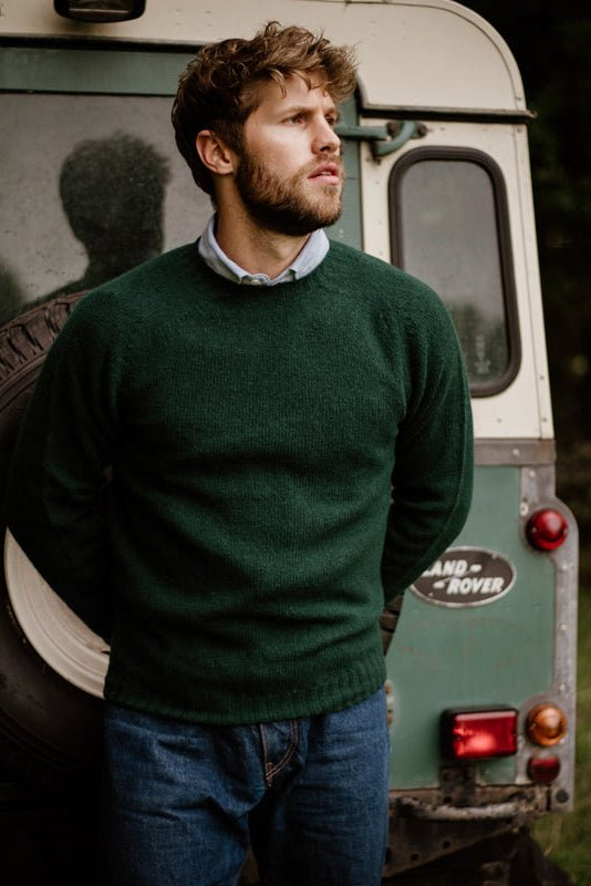 A man with wavy hair and a beard, wearing the Campbells of Beauly Shetland Jumper in green, stands outdoors in front of an old green Land Rover, looking thoughtfully into the distance.