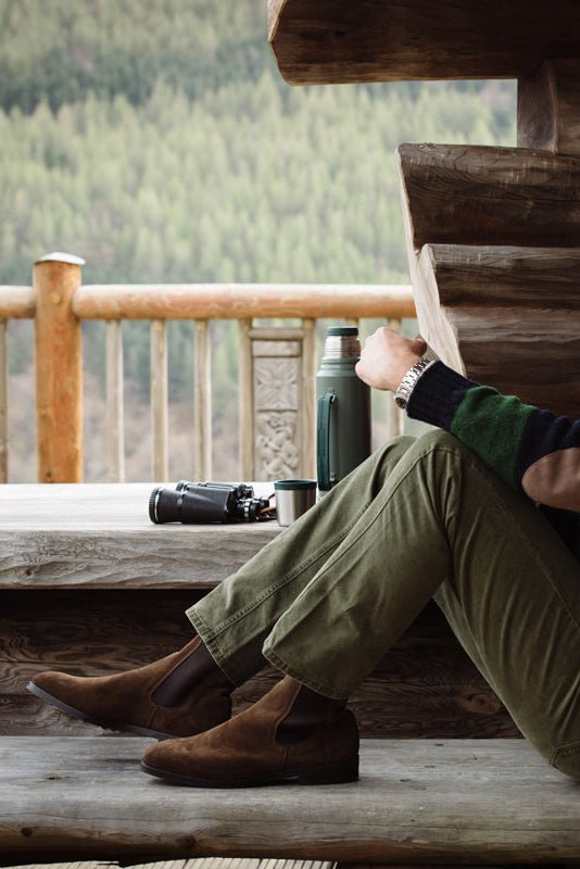 A man reads on a wooden porch in a striped sweater, olive pants, and Suede Chelsea Boots by Campbells of Beauly with Commando soles. A thermos and binoculars sit nearby, set against a forested backdrop.
