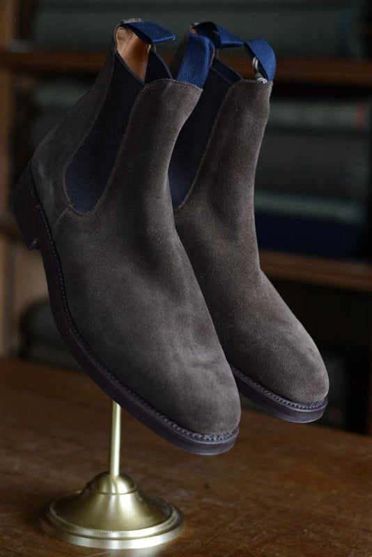 A pair of Campbells of Beauly Suede Chelsea Boots with a Commando sole and navy pull tabs is displayed on a brass stand atop wood, with shelves of folded fabric in the background.