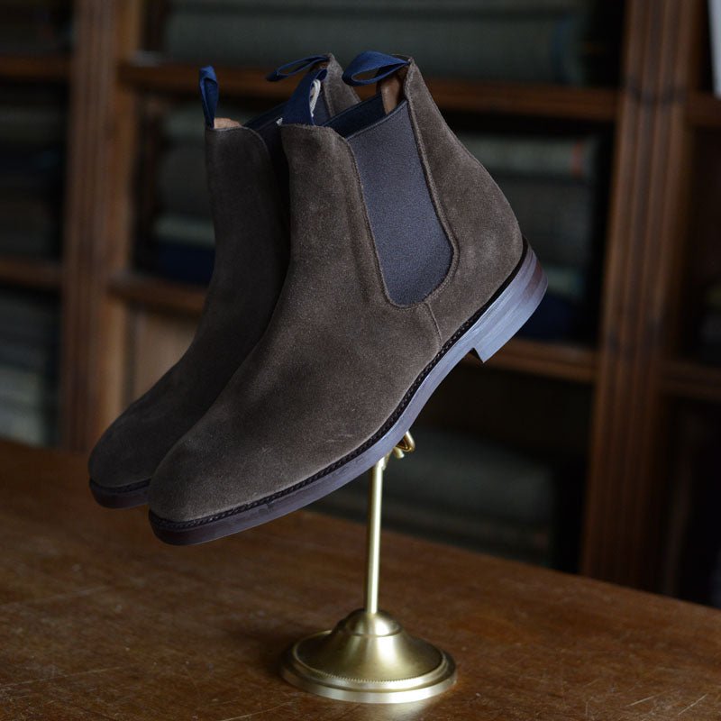 Campbells of Beauly Suede Chelsea Boots in brown, featuring dark elastic side panels and a rugged Commando sole, are showcased on a brass stand atop wood, with shelves of folded fabrics in the background.