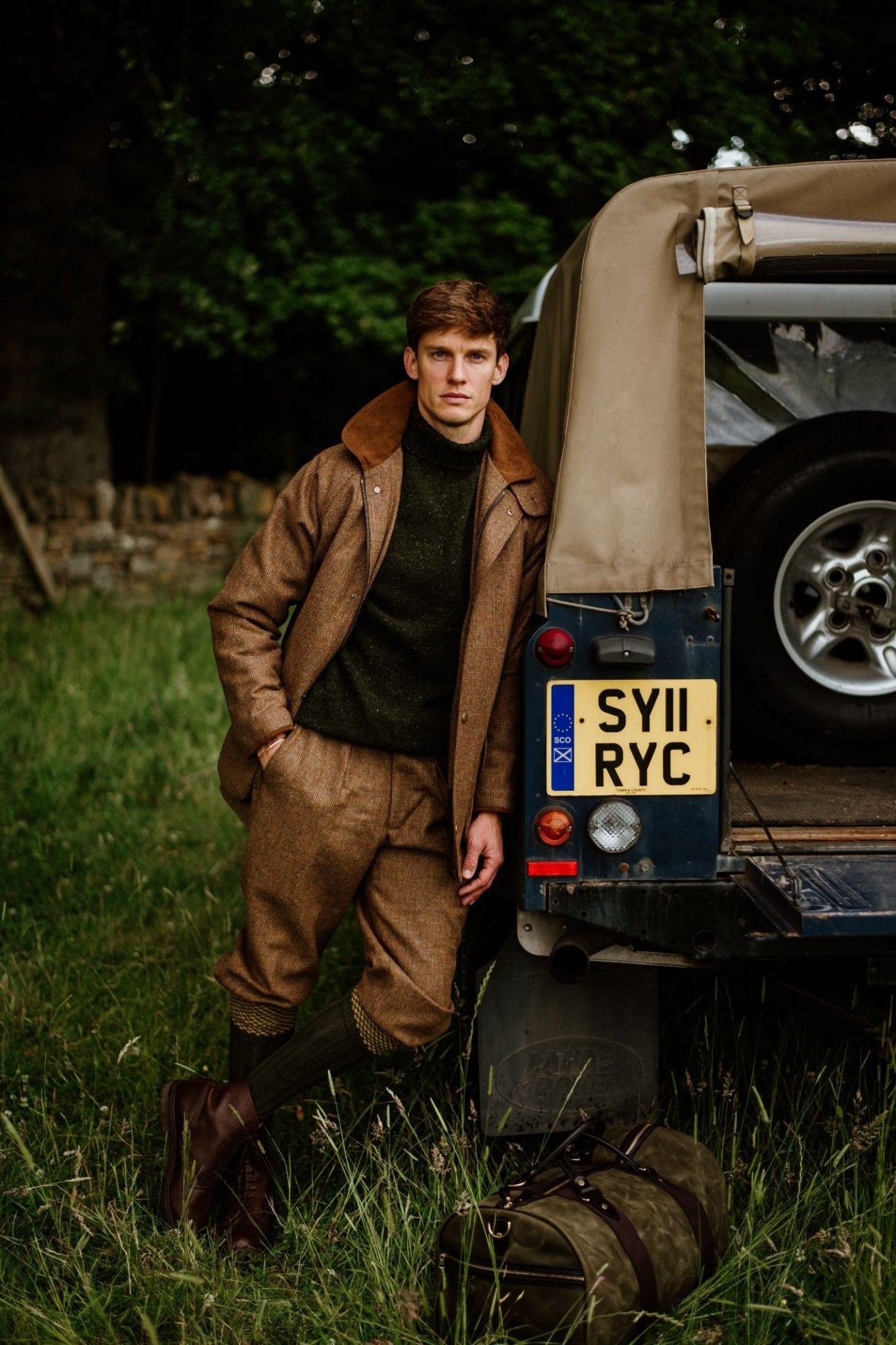 A man wearing Campbells of Beauly Tweed Breeks leans against a blue Land Rover in a grassy field, with trees behind him and a duffel bag on the ground.