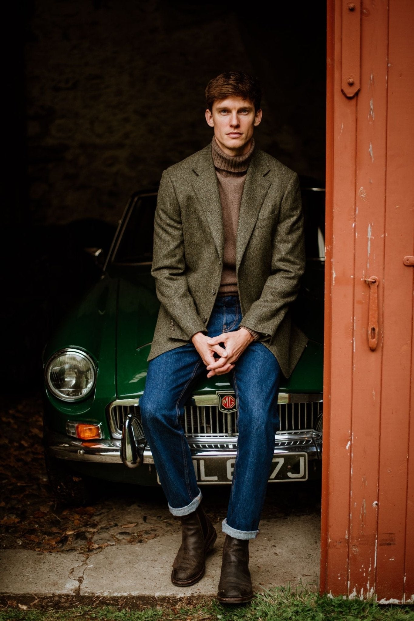 A man in a Campbells of Beauly Forest Herringbone House Jacket sits on the hood of a classic green MG, partially inside a rustic garage with an open red door.