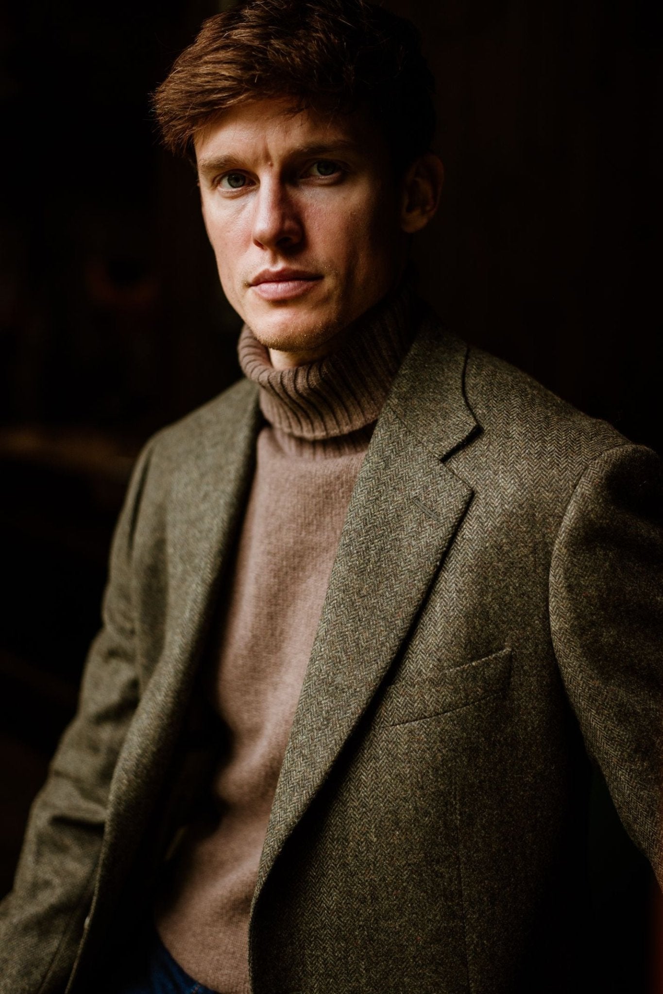 A young man with short brown hair wears a 100% wool tweed turtleneck sweater and the Campbells of Beauly Forest Herringbone House Jacket, looking directly at the camera against a dark background.