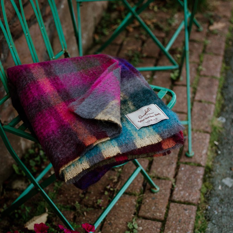 The Campbells of Beauly Painterly Mohair Throw in navy, with its label visible, is draped over a green metal chair on a brick path near a stone wall and plants.