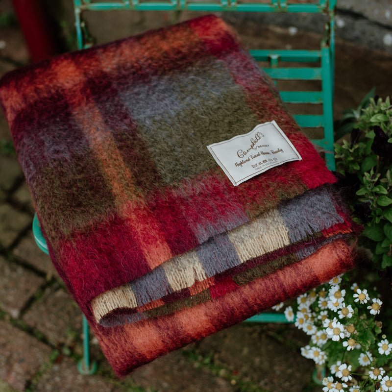 The Painterly Mohair Throw by Campbells of Beauly, in thick rust mohair, is folded on a turquoise metal chair beside small white flowers and greenery. Woven in Yorkshire, its label is sewn onto the soft throw.