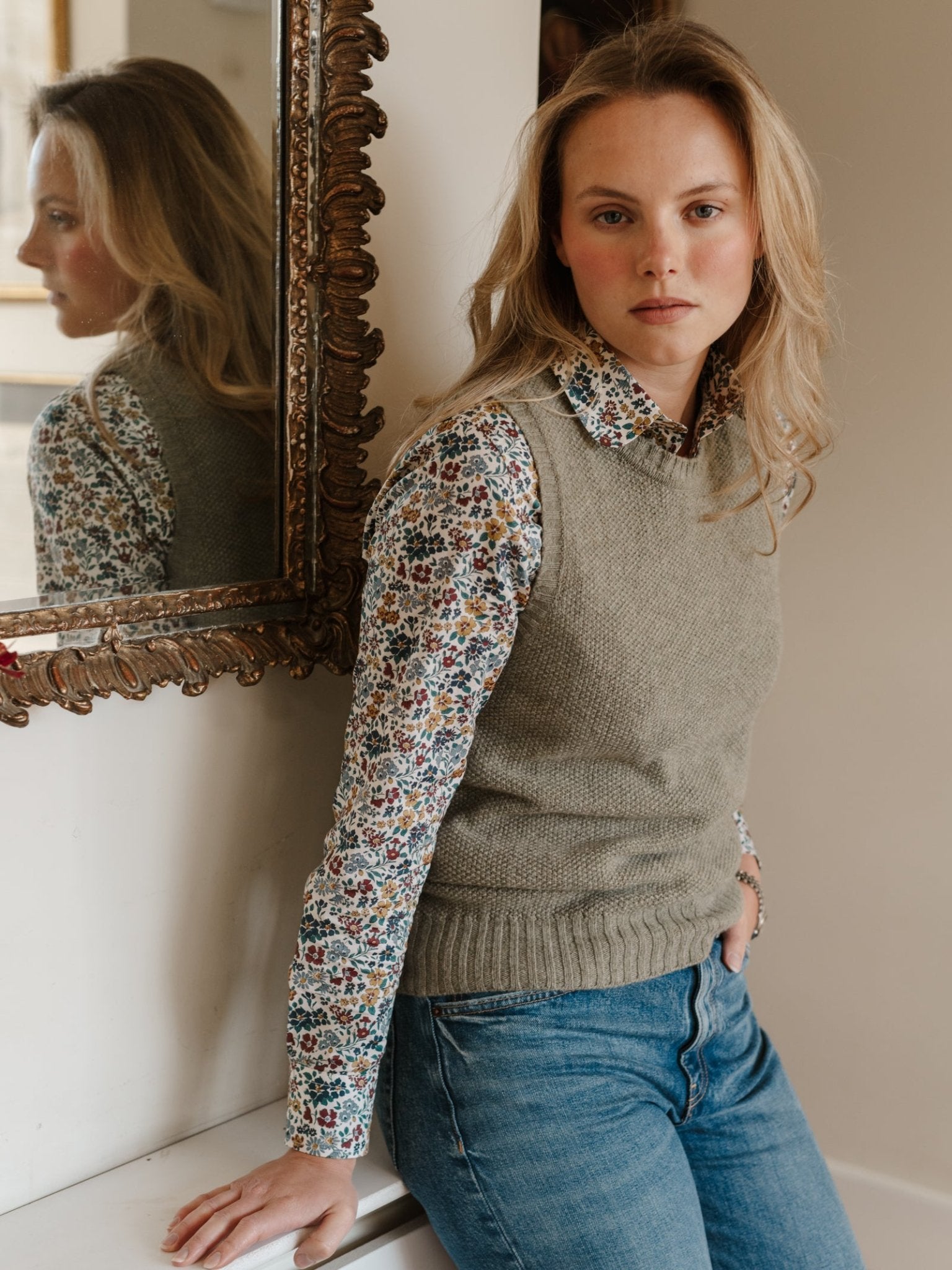 A person with long blonde hair, wearing a floral shirt, Campbell's of Beauly Moss Stitch Crew Tank, and blue jeans sits by an ornate mirror, calmly looking at the camera in a softly lit room.